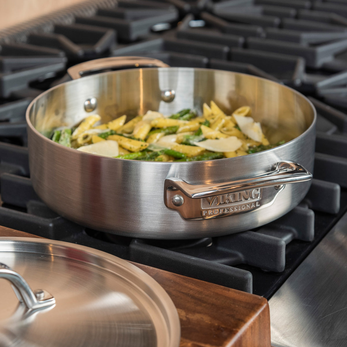 A Viking Culinary Products Professional 5-Ply Stainless Steel 3.4-Quart Casserole Pan with Stay-Cool Handles sits on a gas stove, filled with sliced vegetables, next to its matching lid on a wooden surface.