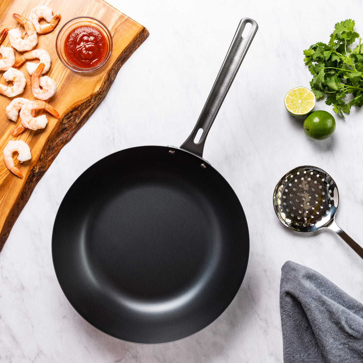 The Viking Blue Carbon Steel 12-Inch Wok by Viking Culinary Products sits on a marble surface with raw shrimp, cocktail sauce on a wooden board, fresh cilantro, a halved lime, a slotted spoon, and a gray towel nearby.