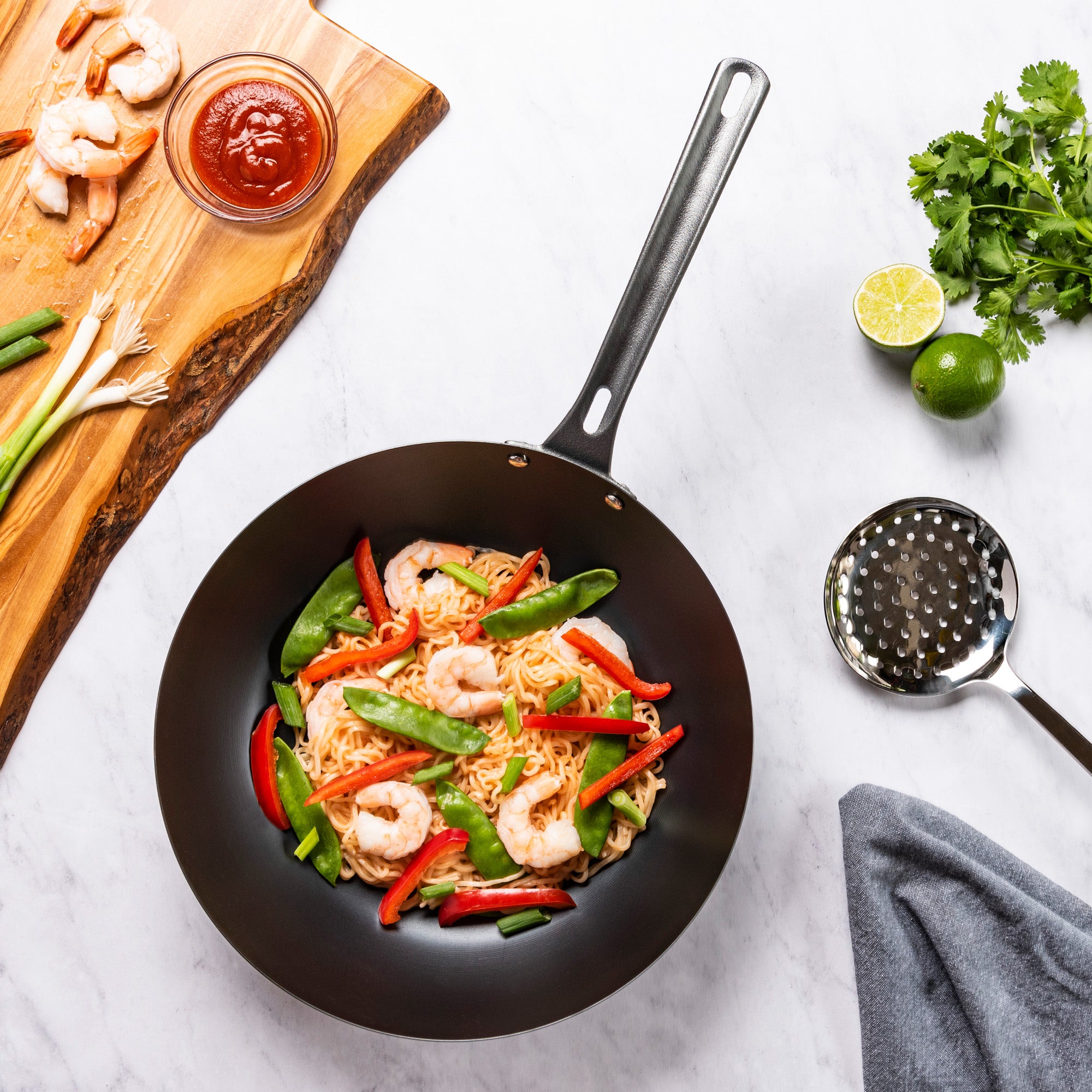 The Viking Culinary Products Viking Blue Carbon Steel 12-Inch Wok holds shrimp, noodles, snap peas, and red bell peppers on a white surface with a nearby board of shrimp, sauce, green onions, cilantro, lime halves, a spatula, and gray cloth.