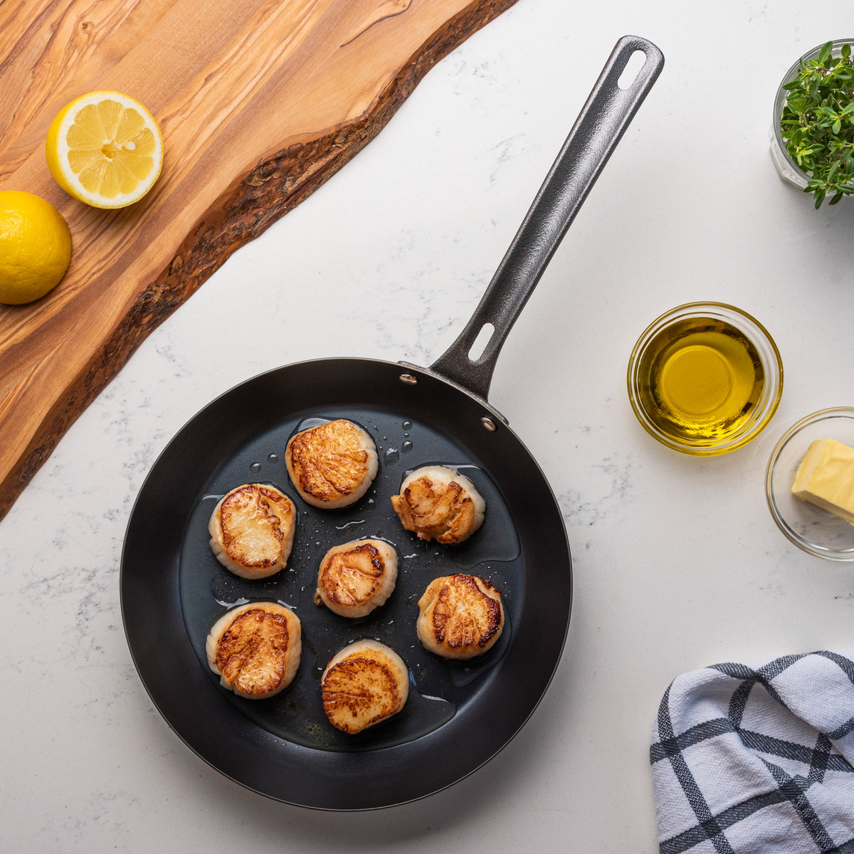 A Viking Culinary Products Viking Blue Carbon Steel 10-Inch Fry Pan with seven seared scallops sits on a white countertop near a halved lemon, olive oil, a pat of butter, a checkered towel, and a partially visible wooden cutting board.