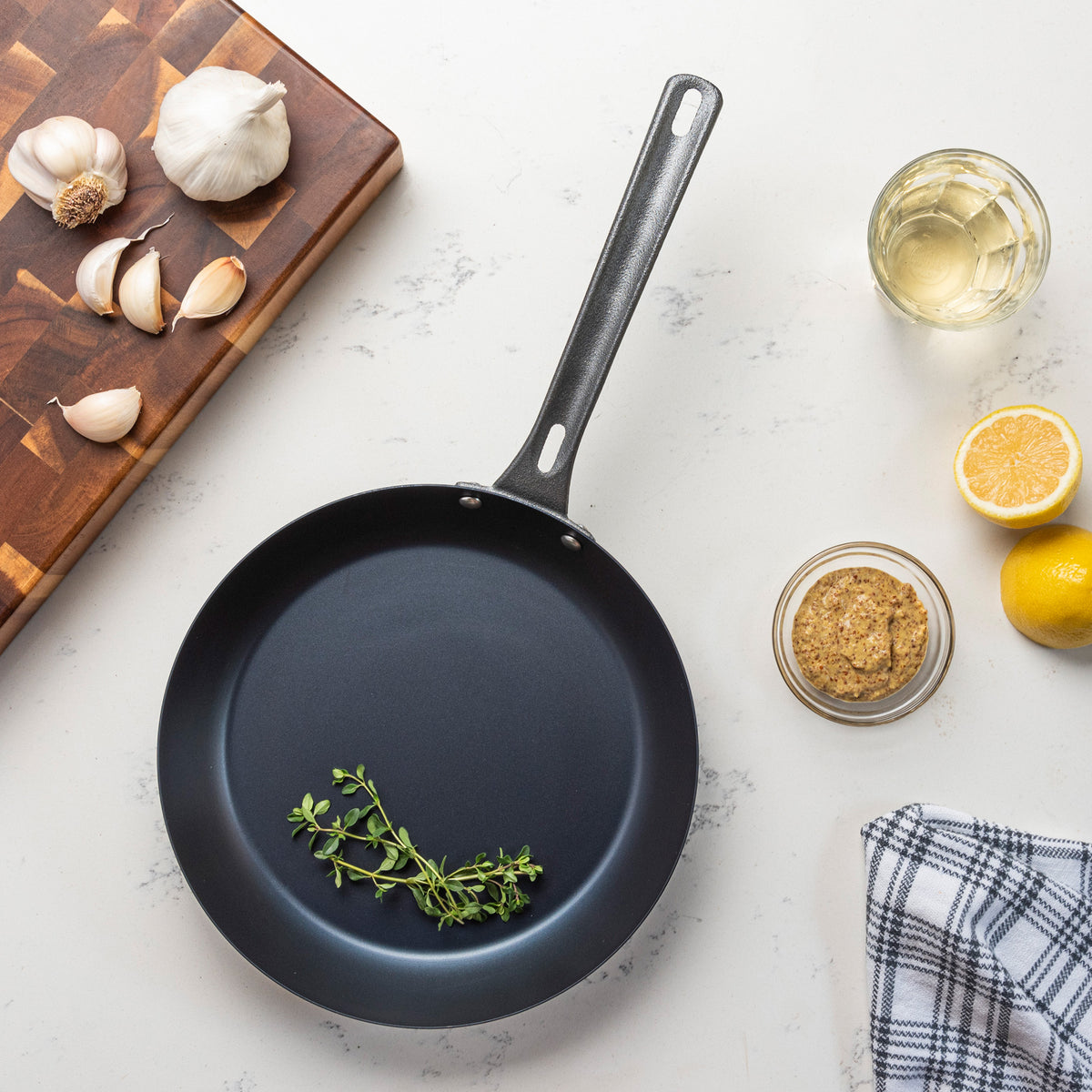 The Viking Blue Carbon Steel 10-Inch Fry Pan by Viking Culinary Products, black in color, holds fresh thyme and sits on a light countertop with garlic, white wine, a halved lemon, mustard, a checkered towel, and a cutting board nearby.