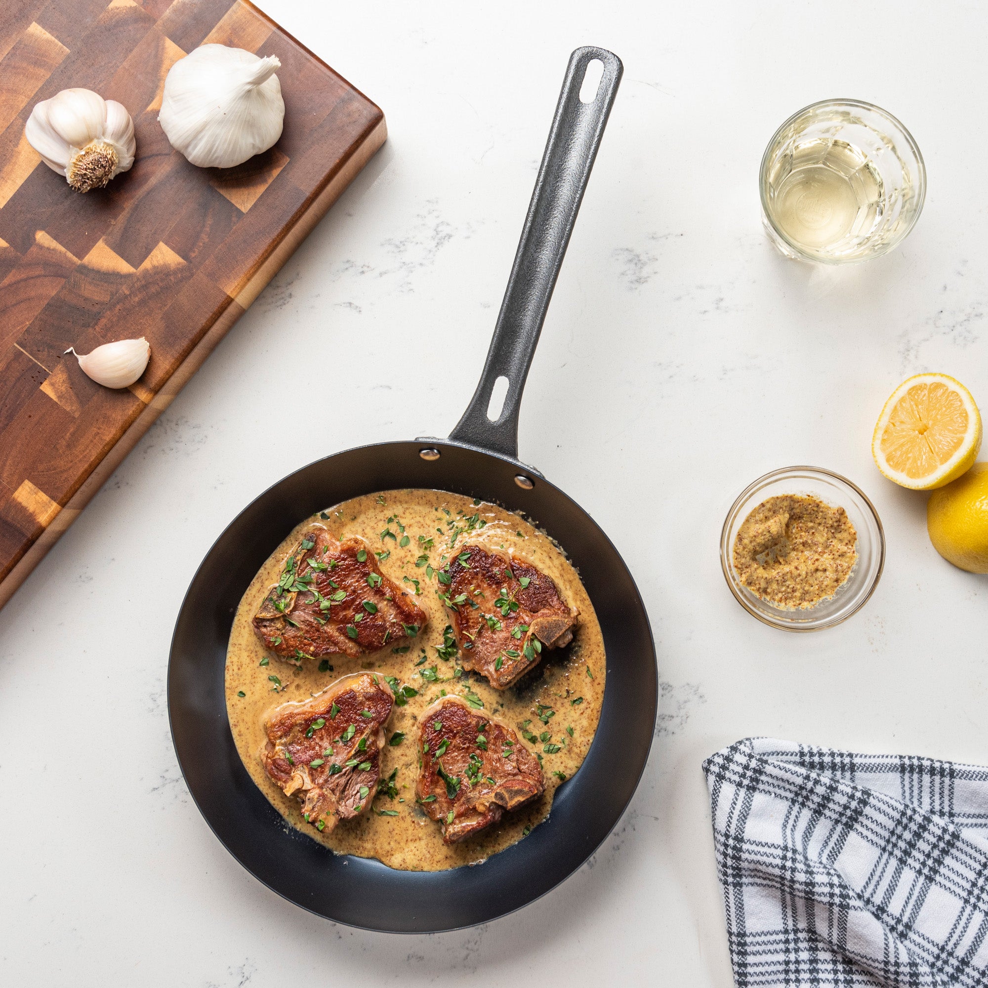 The Viking Blue Carbon Steel 10-Inch Fry Pan by Viking Culinary Products, filled with four pieces of seasoned meat in creamy herb sauce, rests on a white counter near garlic, a cutting board, towel, mustard, lemon, and a glass of white wine.