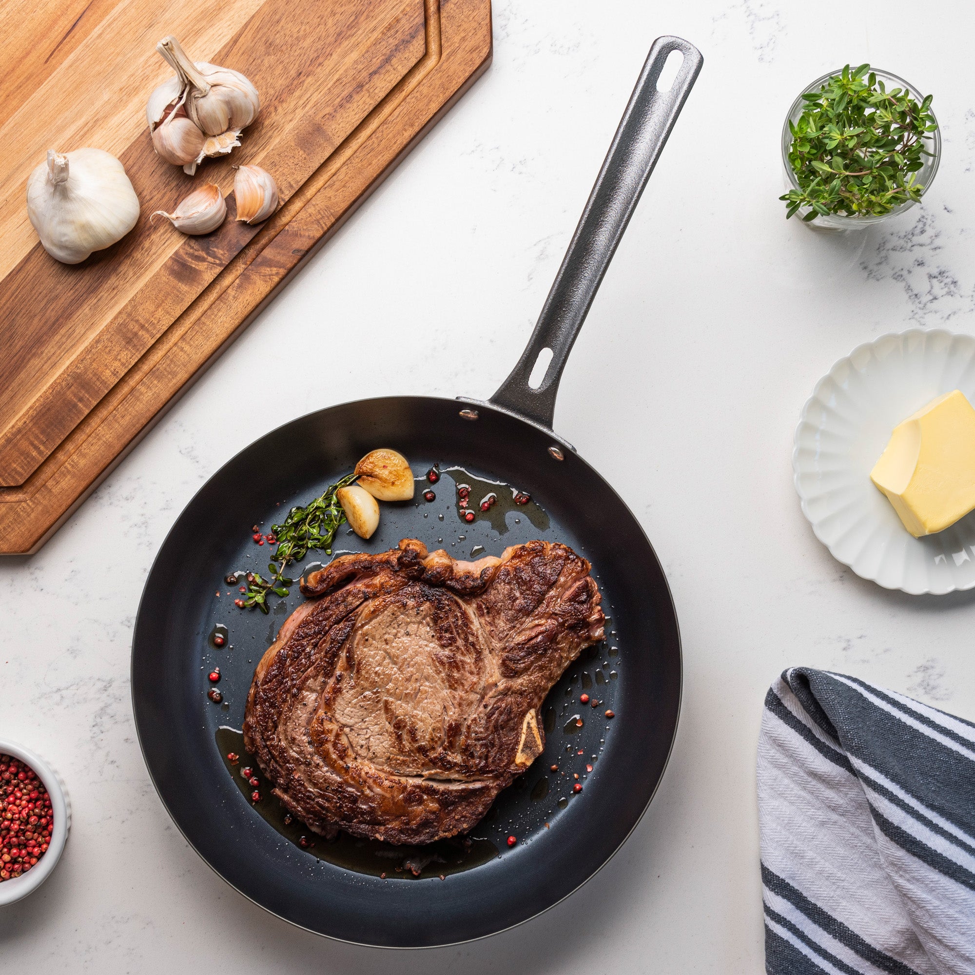 A cooked steak in a Viking Culinary Products Viking Blue Carbon Steel 12-Inch Fry Pan with garlic and herbs, set on a wooden board with garlic, butter, pink peppercorns, fresh herbs, and a striped towel on a white countertop.