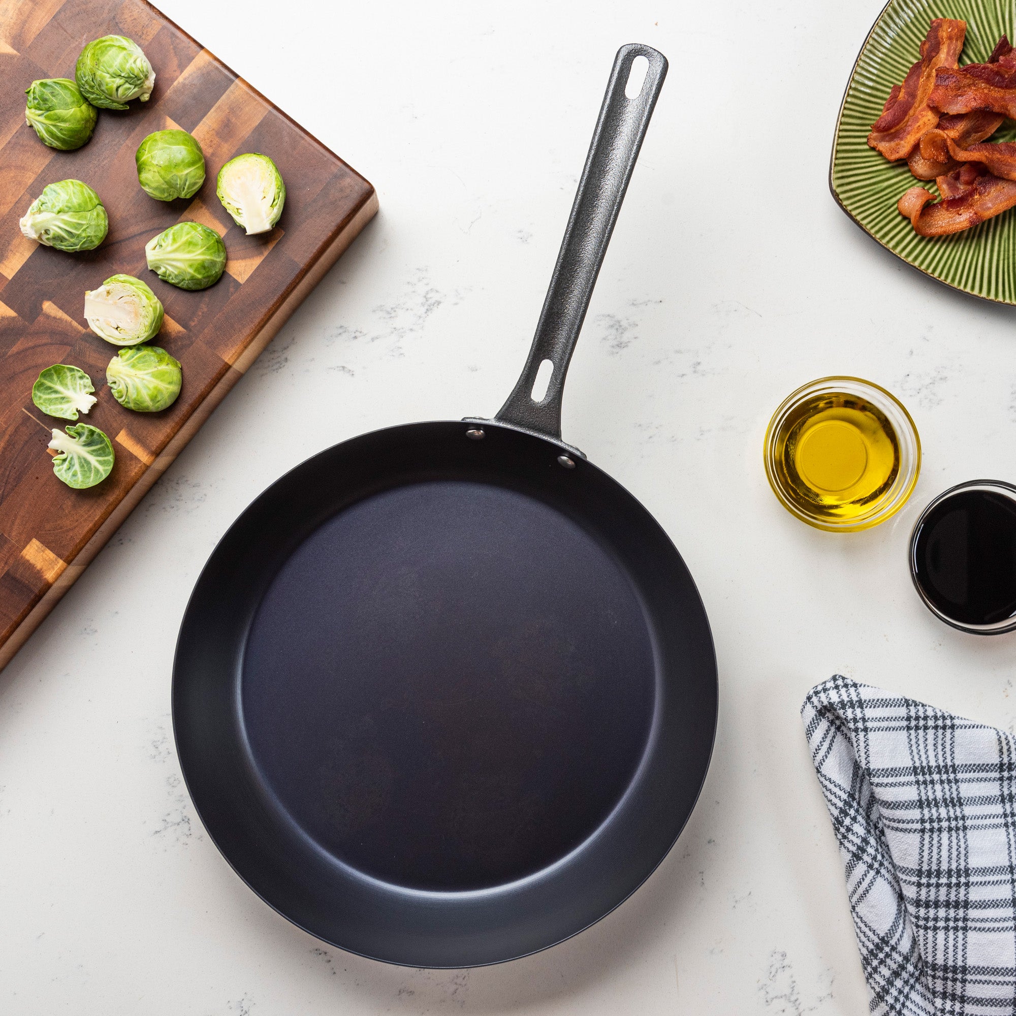 The Viking Culinary Products Blue Carbon Steel 12-Inch Fry Pan rests on a white countertop, surrounded by a cutting board with halved Brussels sprouts, cooked bacon on a plate, bowls of olive oil and balsamic vinegar, and a checkered kitchen towel.