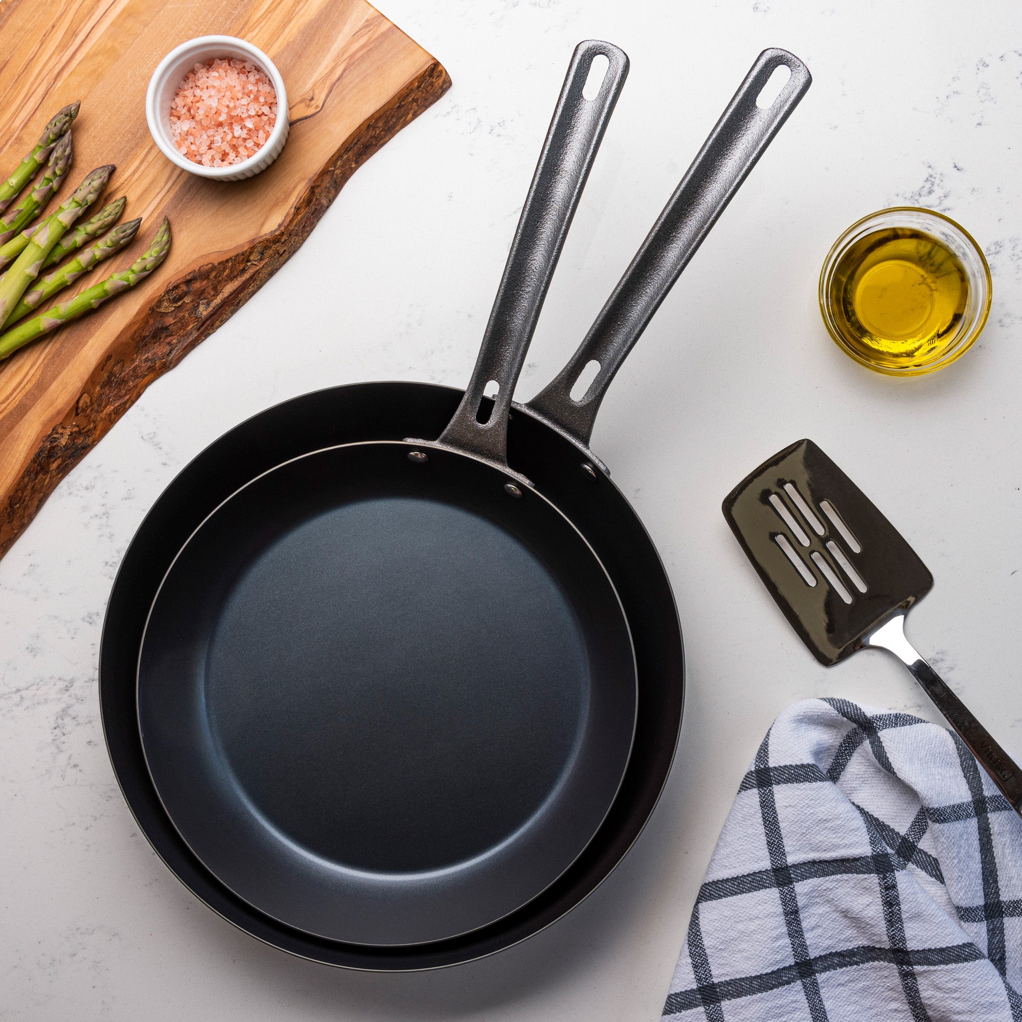 The Viking 2-Piece Blue Carbon Steel Fry Pan Set by Viking Culinary Products sits on a white countertop next to a wooden board with asparagus, pink salt, olive oil, a metal spatula, and a checkered towel—ideal for lightweight cookware lovers.