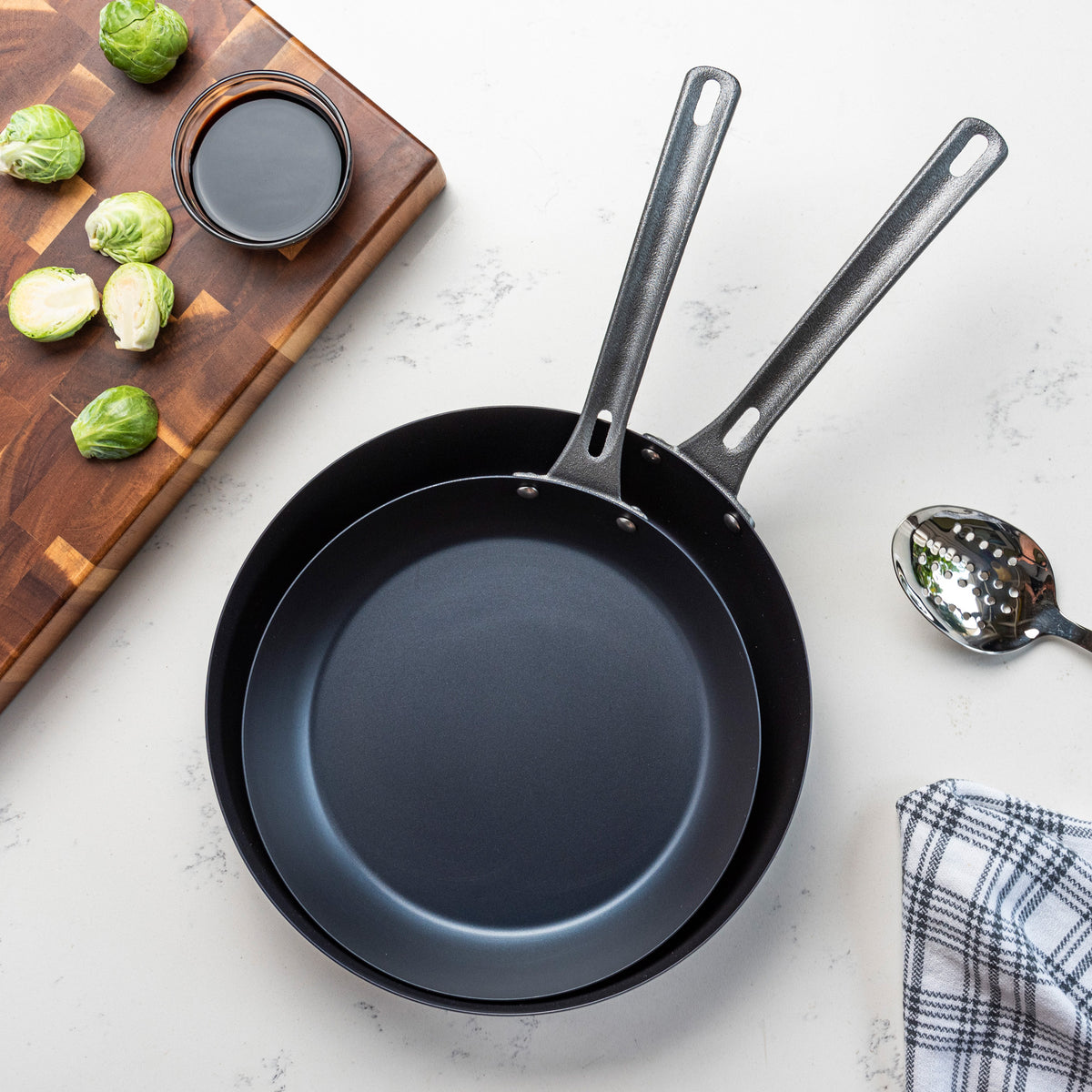 Two empty Viking 2-Piece Blue Carbon Steel Fry Pans by Viking Culinary Products, featuring metal handles, rest on a white countertop beside a slotted spoon, checkered cloth, cutting board with Brussels sprouts, and a small bowl of dark sauce.