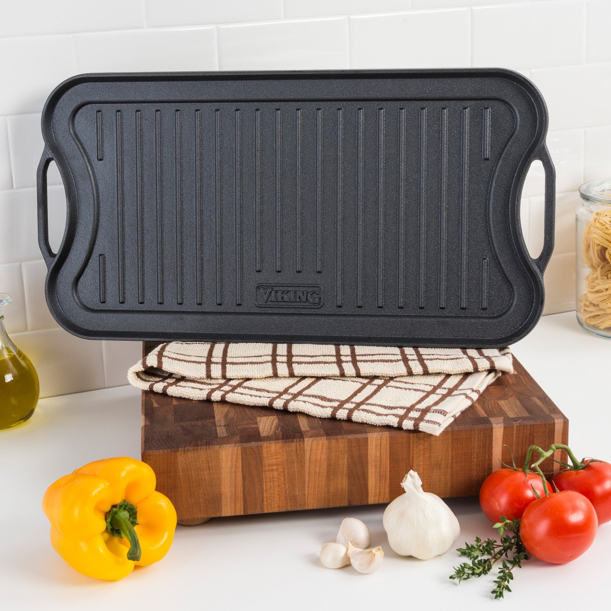 A Viking Culinary Products Cast Iron 20-Inch Reversible Grill/Griddle Pan sits on a folded kitchen towel on a wooden cutting board, surrounded by a yellow bell pepper, garlic, tomatoes, pasta in a jar, and olive oil.