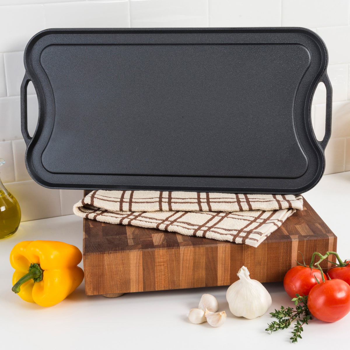 The Viking Cast Iron 20-Inch Reversible Grill/Griddle Pan by Viking Culinary Products, shown upright on a wooden board with plaid towel, is surrounded by tomatoes, garlic, yellow bell pepper, and herbs on a white countertop.