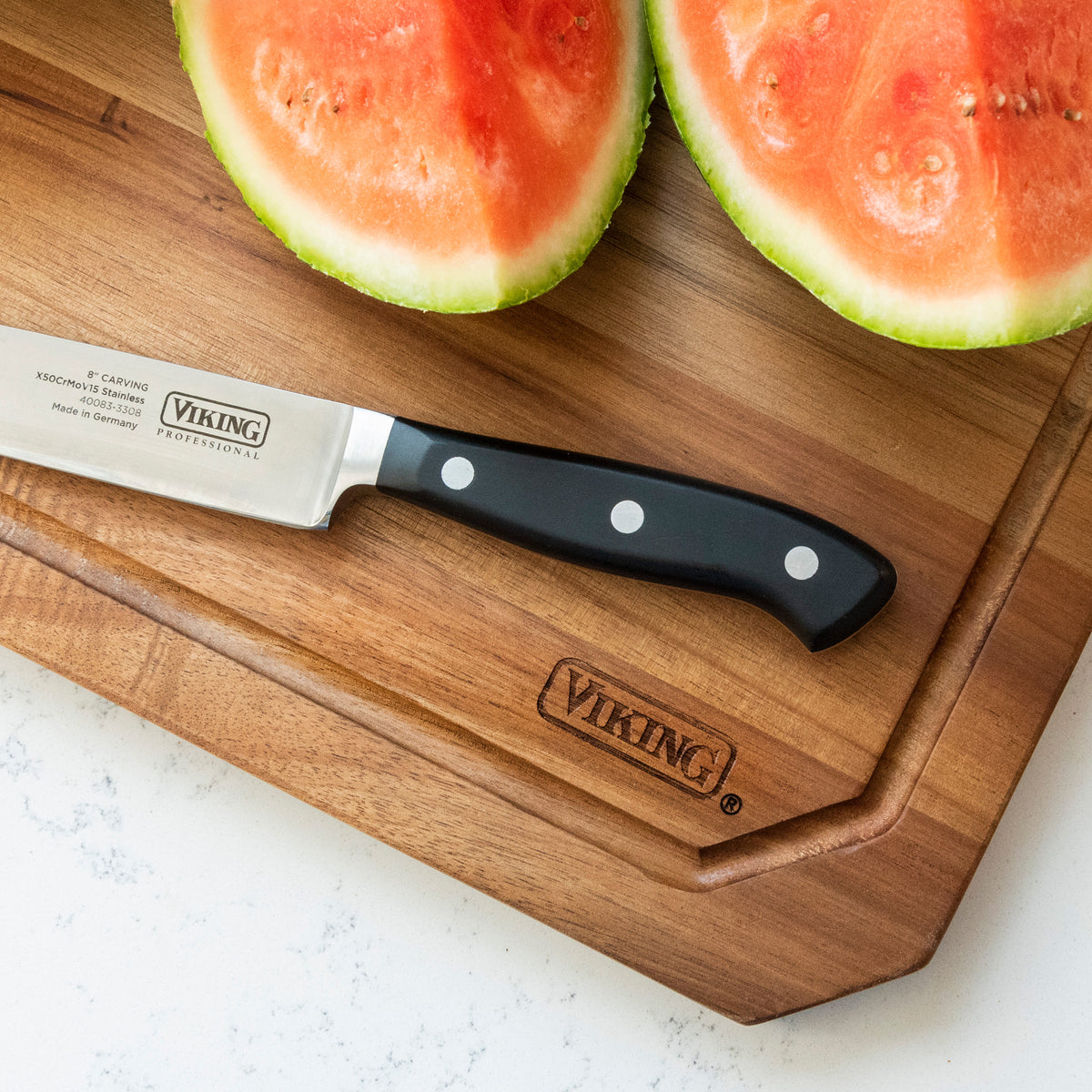 A Viking Acacia Carving Board with Juice Groove by Viking Culinary Products, engraved with the Viking logo, holds a slicing knife with a black riveted handle beside a halved watermelon; the board measures 18 x 12 inches.