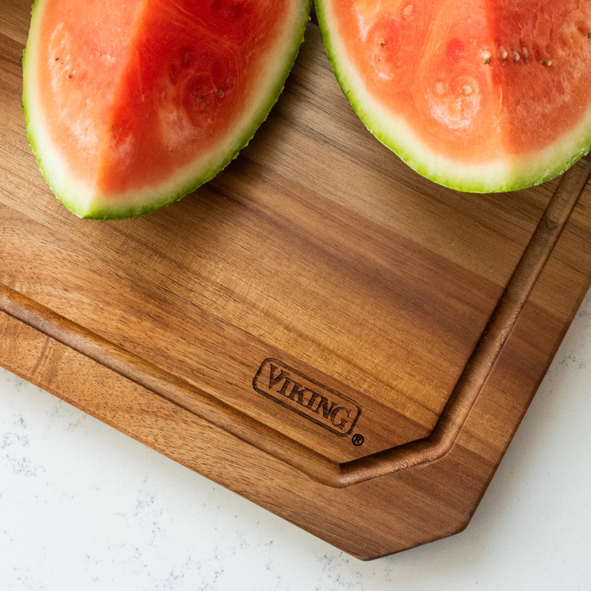 Two slices of watermelon rest on a Viking Culinary Products Acacia Carving Board with Juice Groove, placed on a white, lightly speckled countertop.