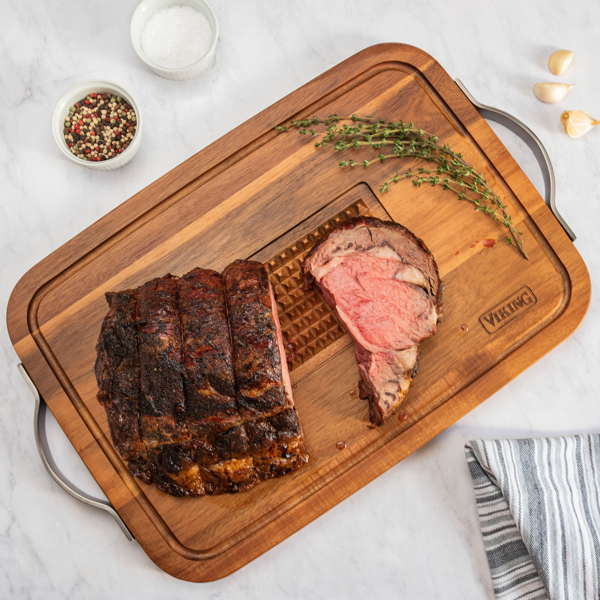 Sliced roasted beef on the Viking Acacia Carving Board with Juice Well and Metal Handle by Viking Culinary Products, garnished with thyme, alongside bowls of salt, peppercorns, garlic cloves, and a striped napkin on a marble countertop.