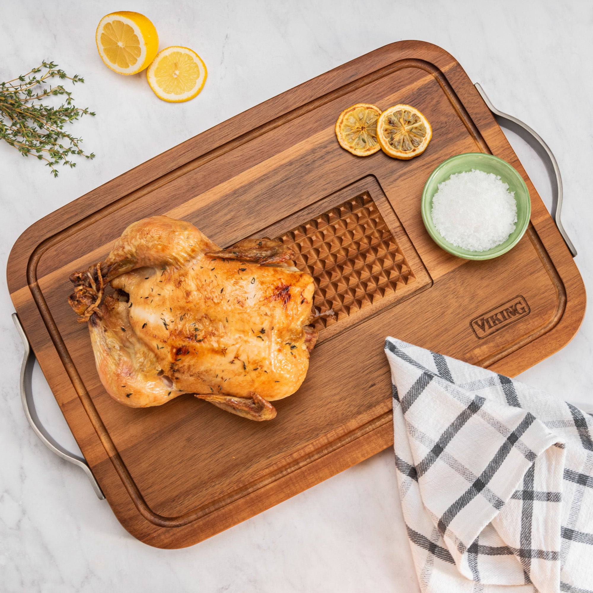 A whole roasted chicken on the Viking Acacia Carving Board with Juice Well and Metal Handle by Viking Culinary Products, alongside a green bowl of salt, a checkered towel, lemon slices, and fresh thyme on a white surface.
