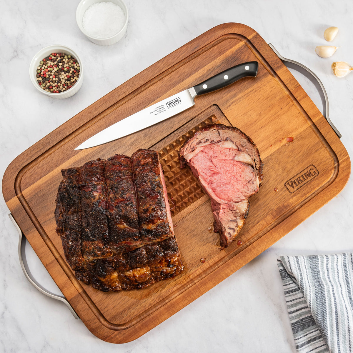 The Viking Acacia Carving Board with Juice Well and Metal Handle by Viking Culinary Products holds sliced roast beef, a carving knife, and seasonings. Crafted from acacia wood, it features heavy-duty metal handles and a drip-catching juice well.