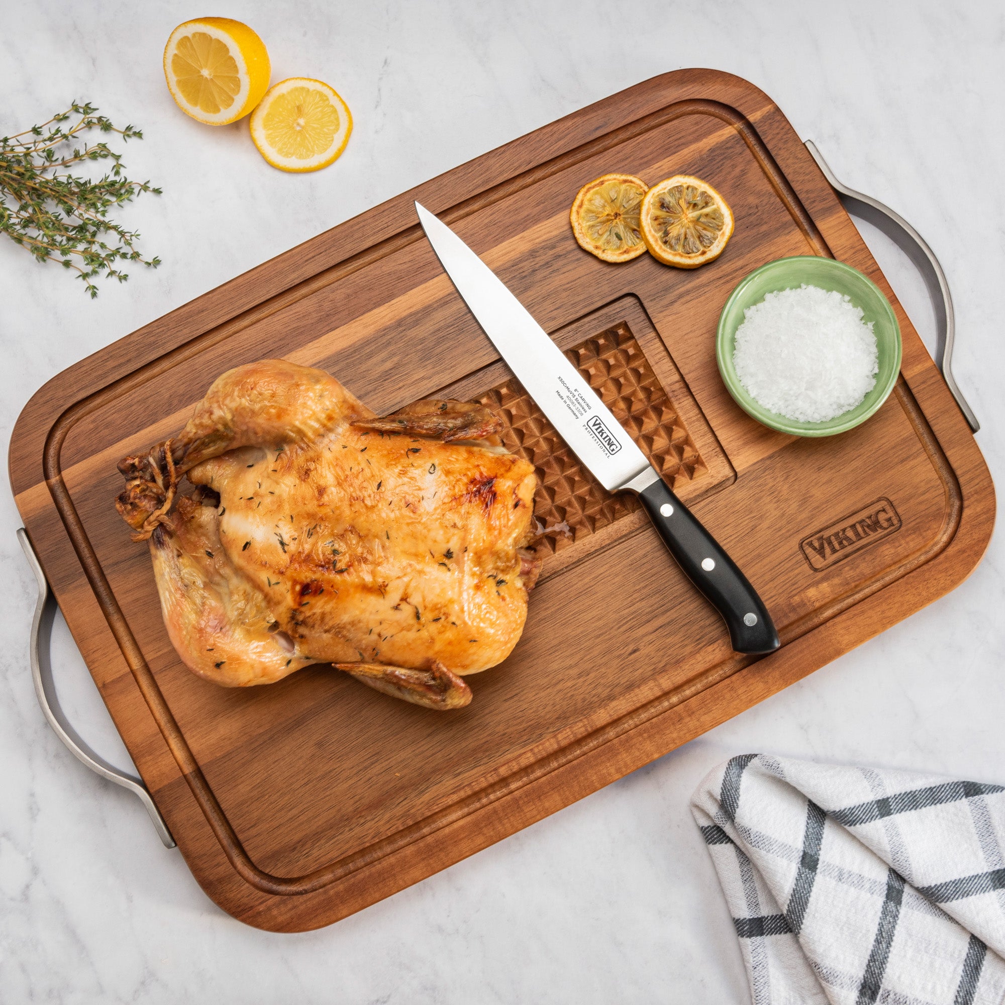 Roast chicken on the Viking Acacia Carving Board with Juice Well and Metal Handle by Viking Culinary Products, with a carving knife, sea salt dish, lemon slices, fresh thyme, and a blue-and-white towel on a marble countertop.