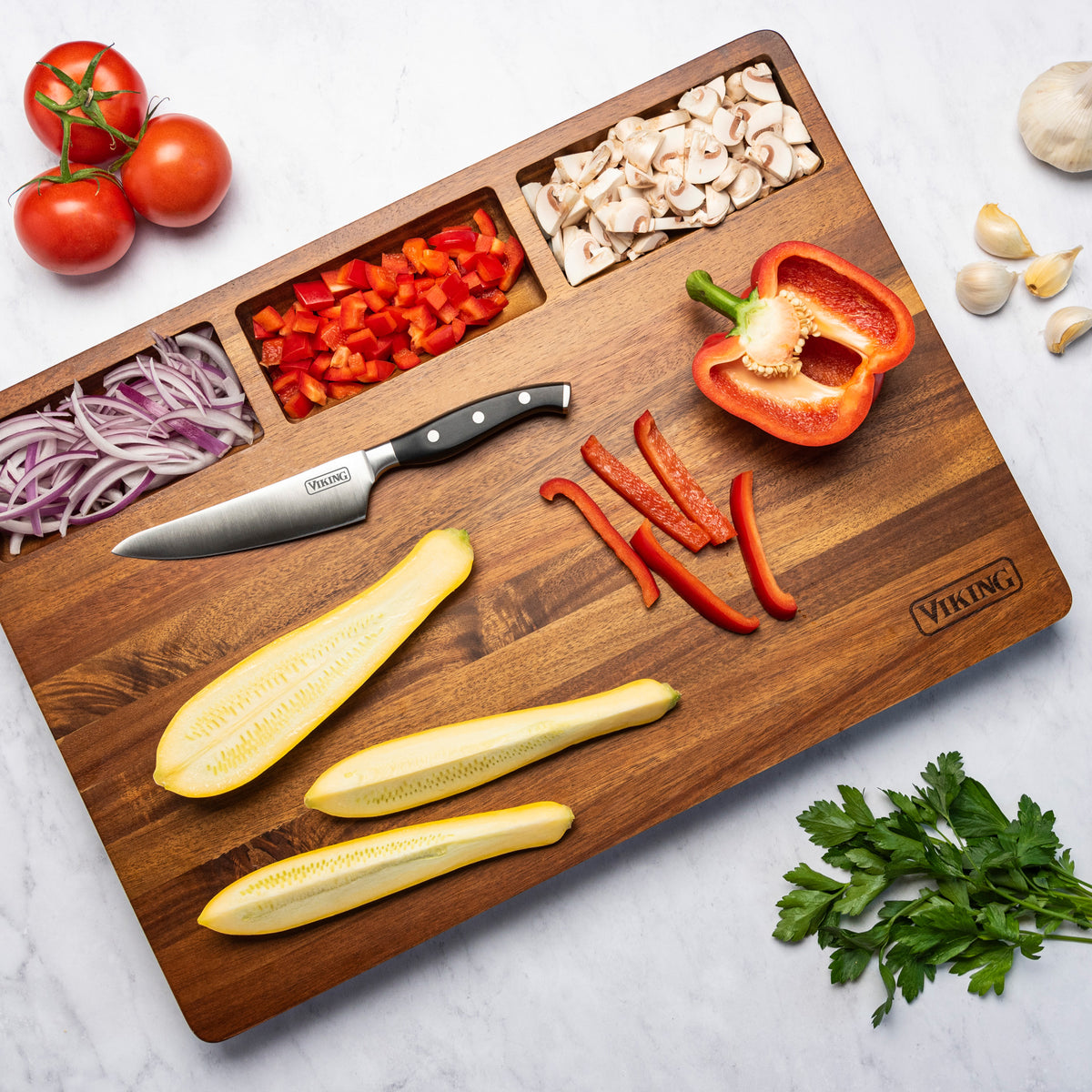 The Viking Acacia 20-Inch Reversible Butcher Block Prep and Carving Board by Viking Culinary Products features compartments holding chopped red onion, bell pepper, and mushrooms, plus sliced zucchini, a knife, whole veggies nearby, and a built-in juice groove.