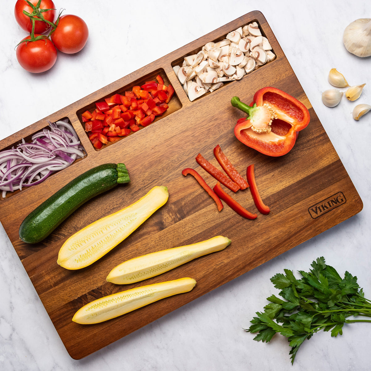 The Viking Acacia 20-Inch Reversible Butcher Block Prep and Carving Board by Viking Culinary Products holds chopped red bell pepper, mushrooms, and onion in built-in sections, with sliced zucchini, squash, and whole veggies arranged around it on marble.