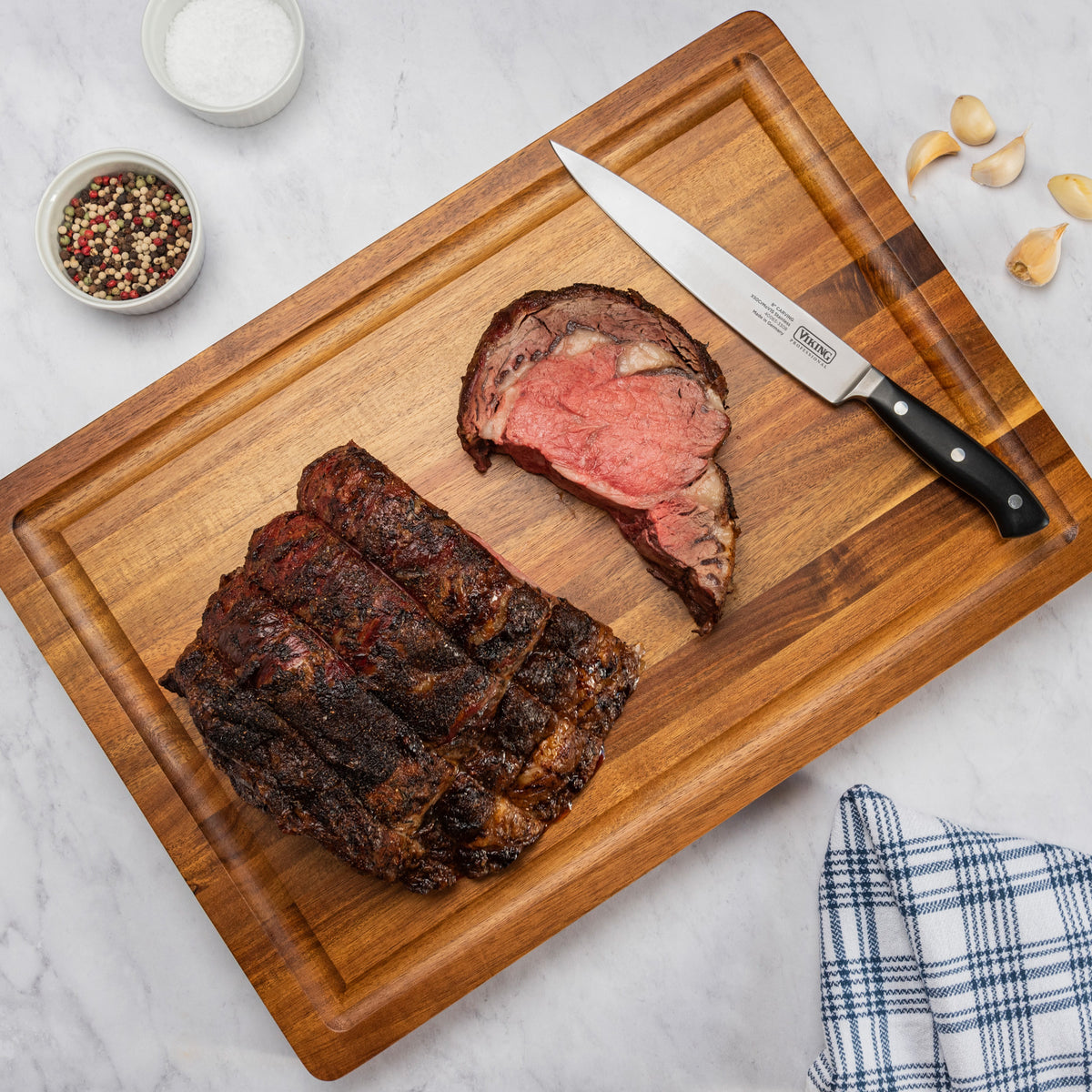 A cooked prime rib roast and sliced piece rest on the Viking Acacia 20-Inch Reversible Butcher Block Prep and Carving Board by Viking Culinary Products, with a knife, garlic cloves, peppercorns, salt, and a blue-checked towel on a marble countertop.