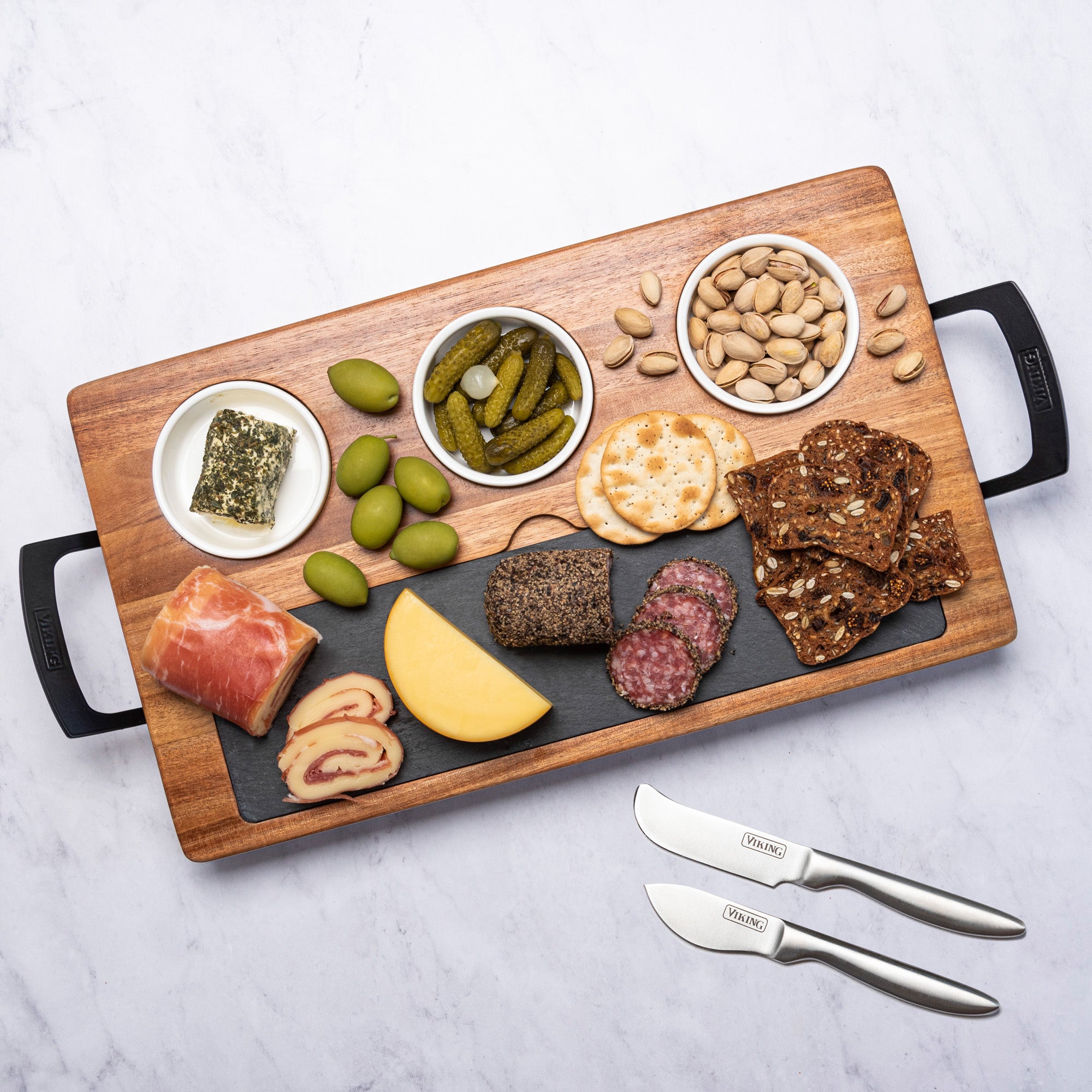 The Viking Culinary Products Viking 7-Piece Acacia Wood Slate Cheese Board Set features an Acacia board with black handles, a slate insert, small bowls for snacks, and two cheese knives, pictured with cheeses, crackers, olives, meats, and nuts.