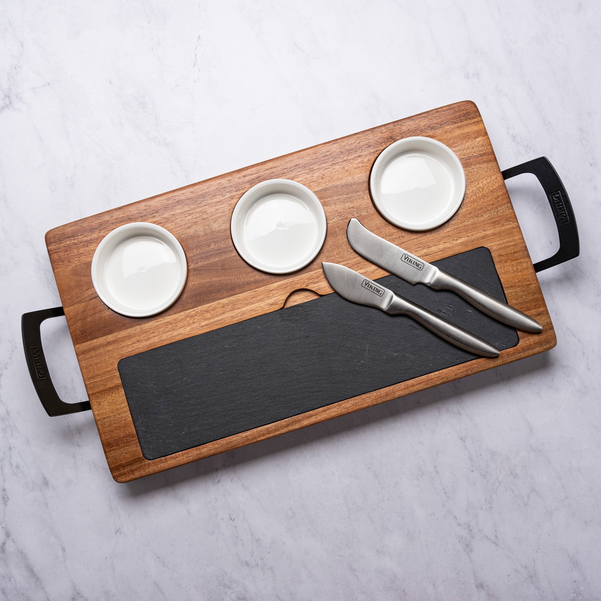 The Viking 7-Piece Acacia Wood Slate Cheese Board Set by Viking Culinary Products includes an acacia board with black handles, three white ceramic ramekins, an inset slate slab, and two stainless steel cheese knives.