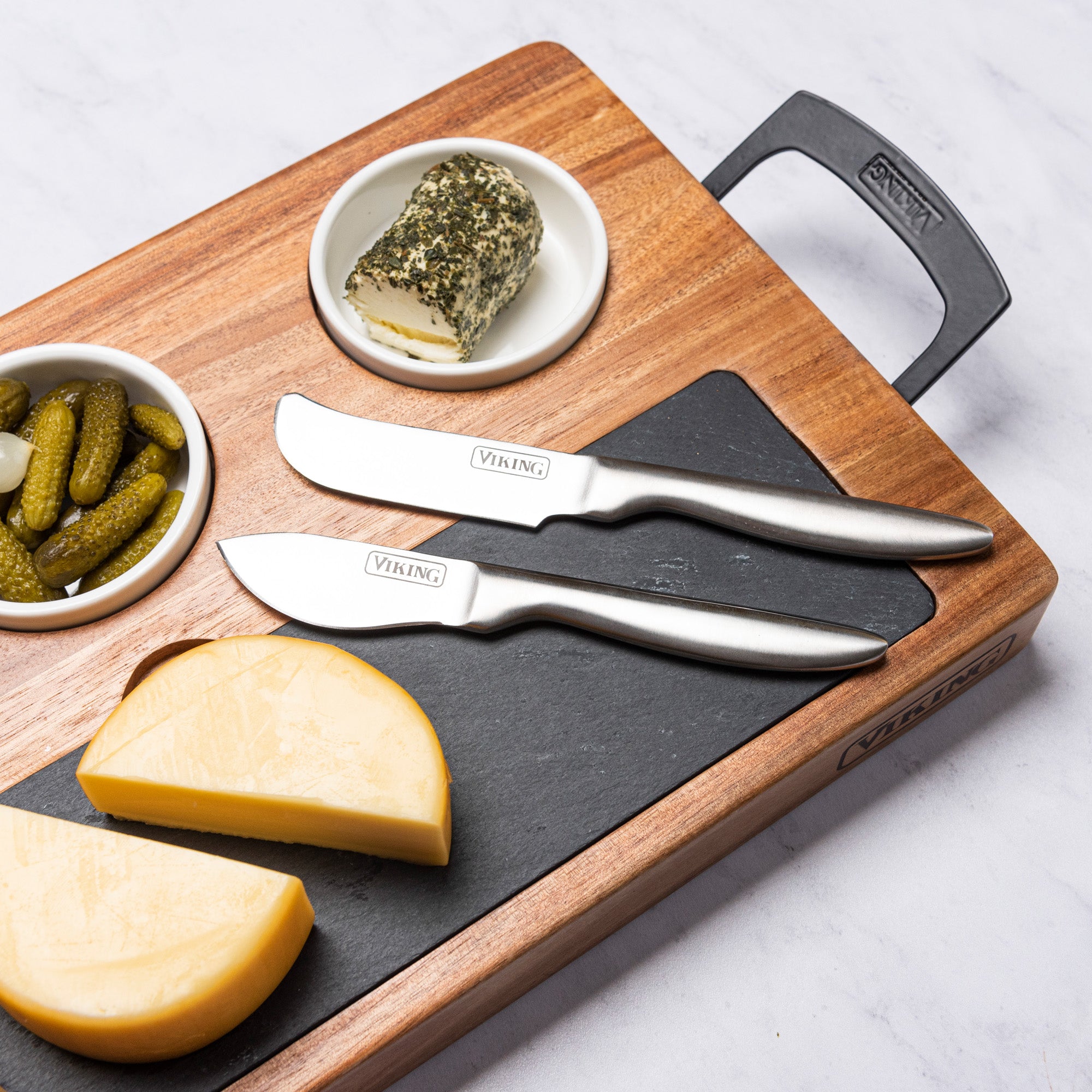 The Viking 7-Piece Acacia Wood Slate Cheese Board Set by Viking Culinary Products displays cheese, pickles, herb-coated goat cheese, and two included cheese knives on a white marble surface.
