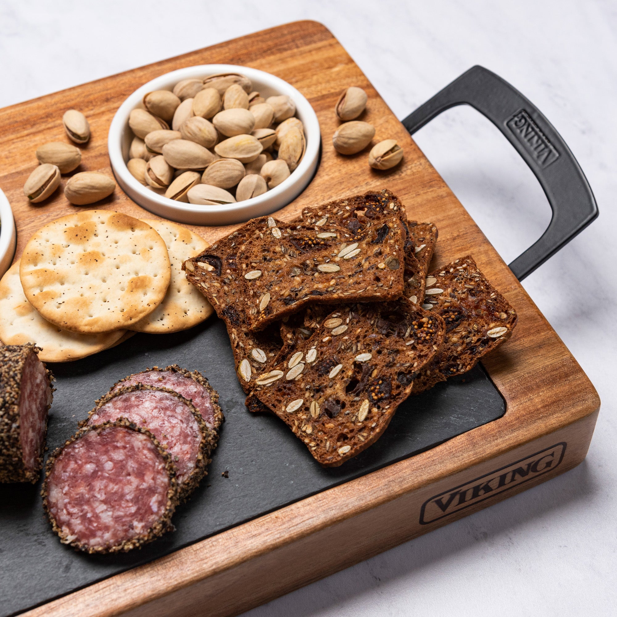 The Viking 7-Piece Acacia Wood Slate Cheese Board Set by Viking Culinary Products holds sliced salami, crackers, seeded crispbread, and pistachios in a small bowl. The branded board features a black handle and rests on a white surface.