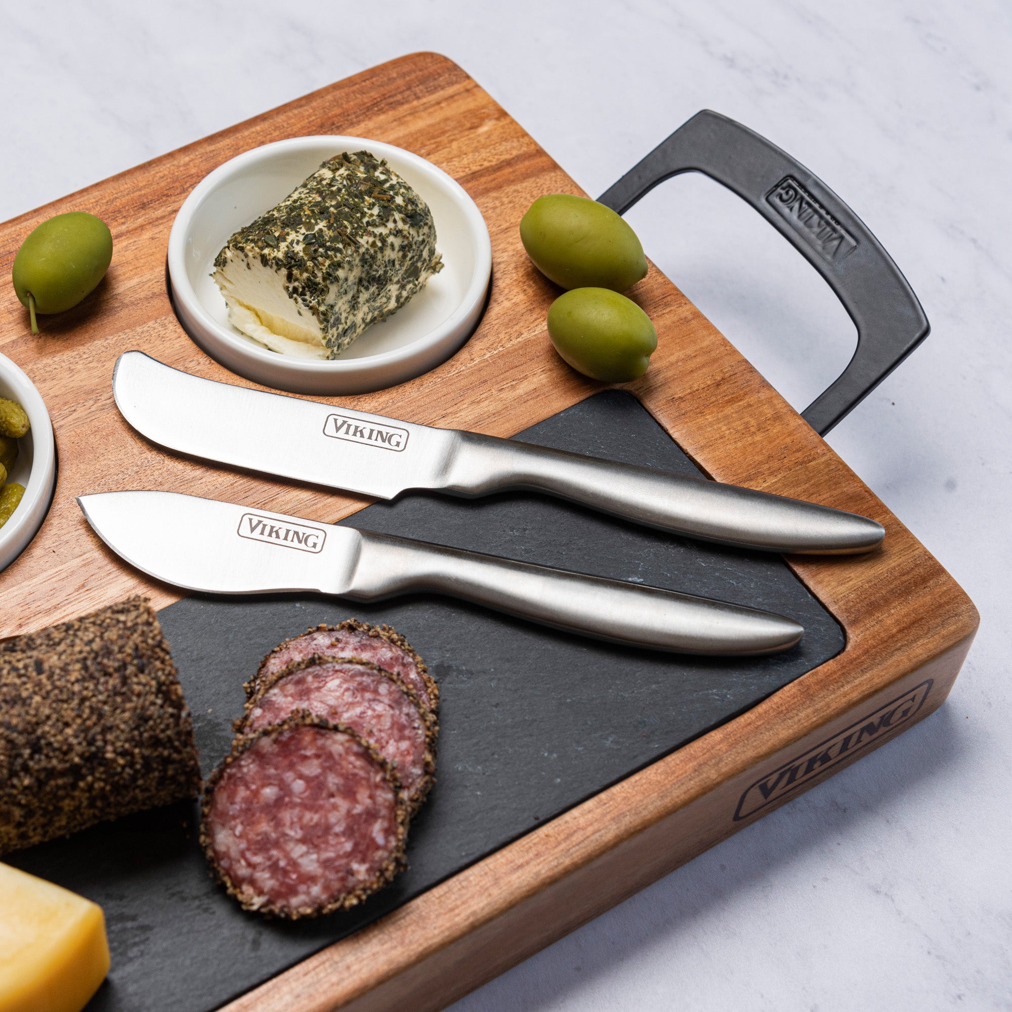 The Viking 7-Piece Acacia Wood Slate Cheese Board Set by Viking Culinary Products holds sliced salami, cheese, herbed cheese dip, green olives, and includes two Viking-labeled cheese knives.