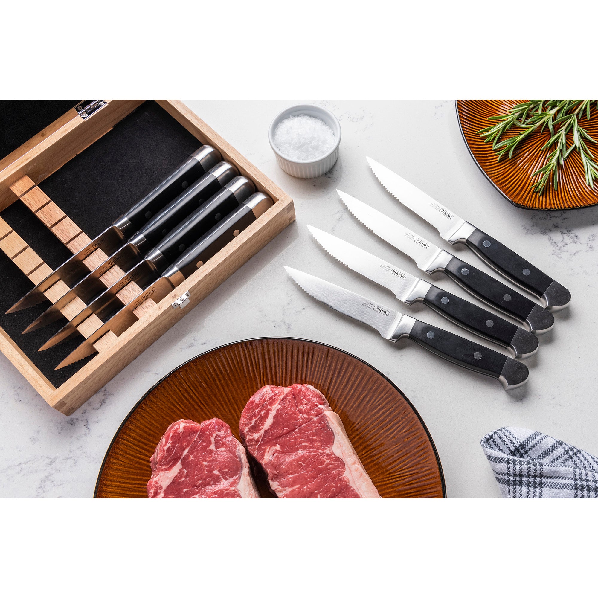 The Viking Steakhouse 8-Piece Steak Knife Set by Viking Culinary Products features corrosion-resistant German stainless steel blades and black double-riveted handles, presented with a gift box on a countertop with steaks, salt, rosemary, and a checked towel.