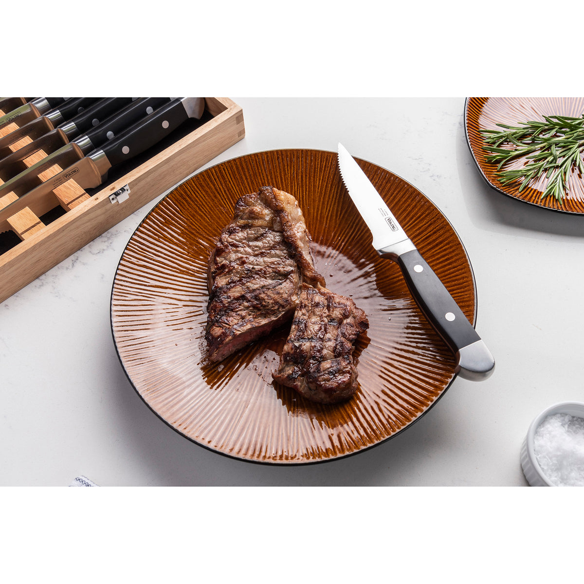 A grilled steak is sliced on a brown plate with a Viking Steakhouse knife by Viking Culinary Products. Beside it is the corrosion-resistant, German steel blade; in the background are the 8-piece gift box, rosemary plate, and salt bowl.