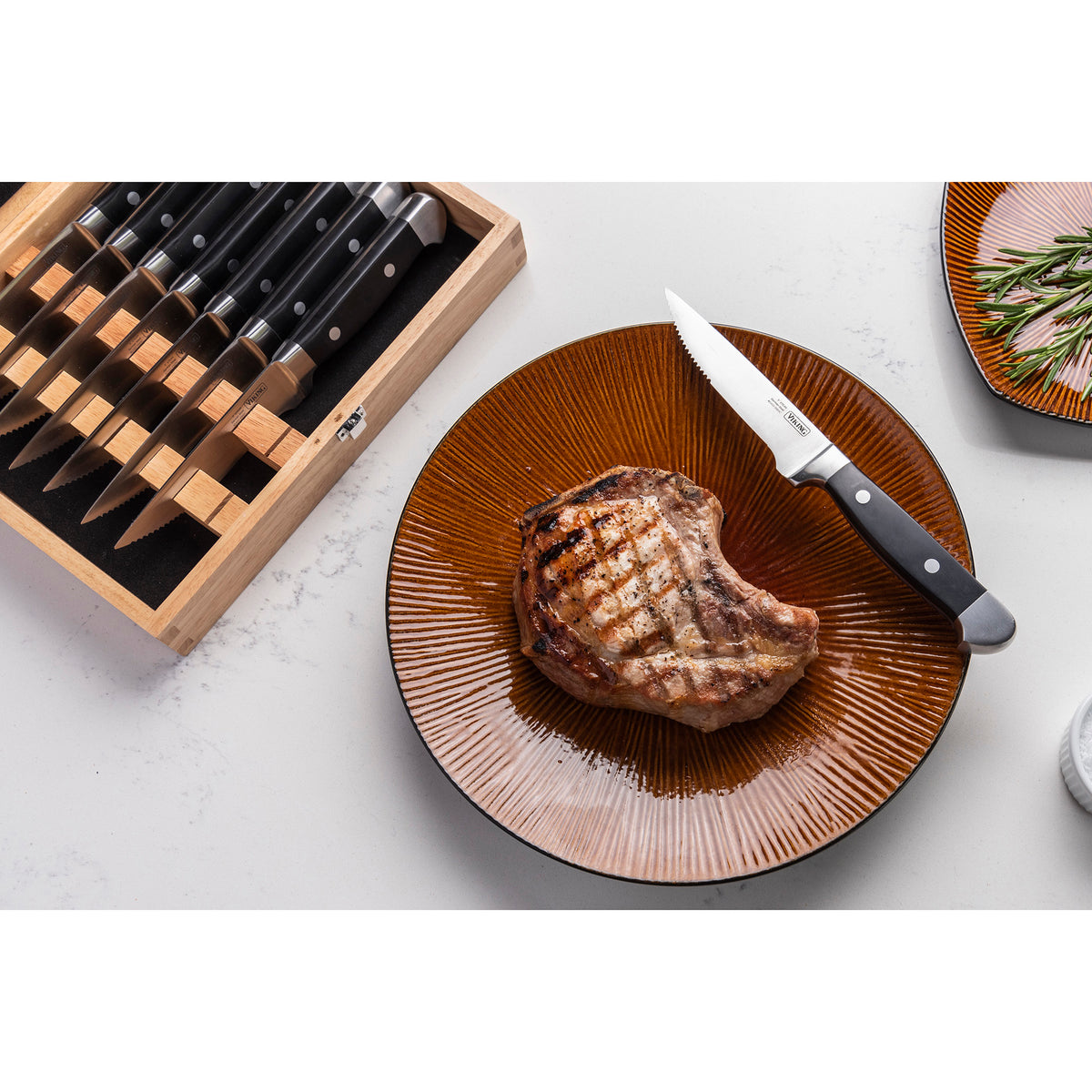 A grilled steak rests on a brown plate beside a Viking Steakhouse 8-Piece Steak Knife from Viking Culinary Products. To the left, the open gift box displays several double-riveted knives. A small plate of herbs appears in the top right corner.