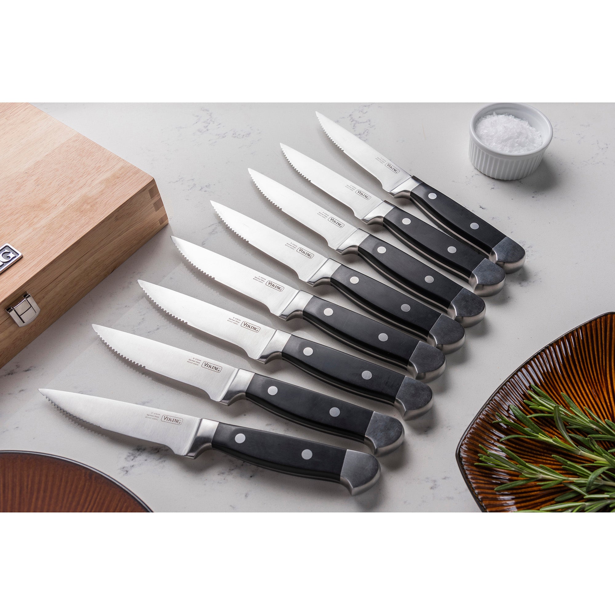 The Viking Steakhouse 8-Piece Steak Knife Set by Viking Culinary Products features corrosion-resistant German stainless steel blades and black double-riveted handles, neatly arranged with a wooden gift box, salt bowl, rosemary sprigs, and a brown plate.