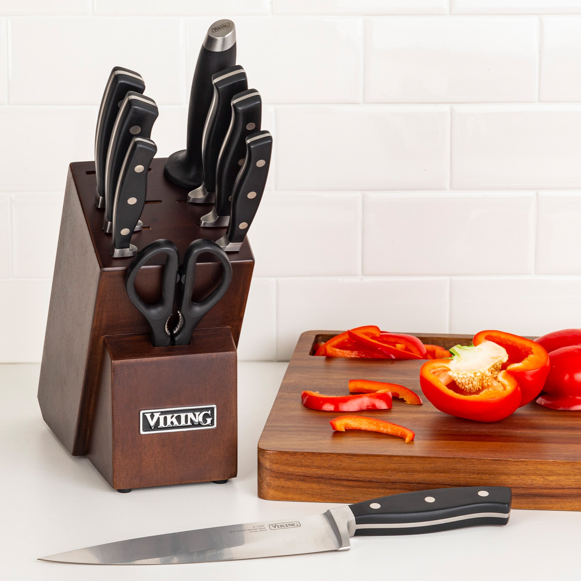 A Viking 10-Piece True Forged Cutlery Set with Block from Viking Culinary Products sits on the counter. A cutting board displays sliced red bell pepper and a chef’s knife, set against a white tile backsplash.