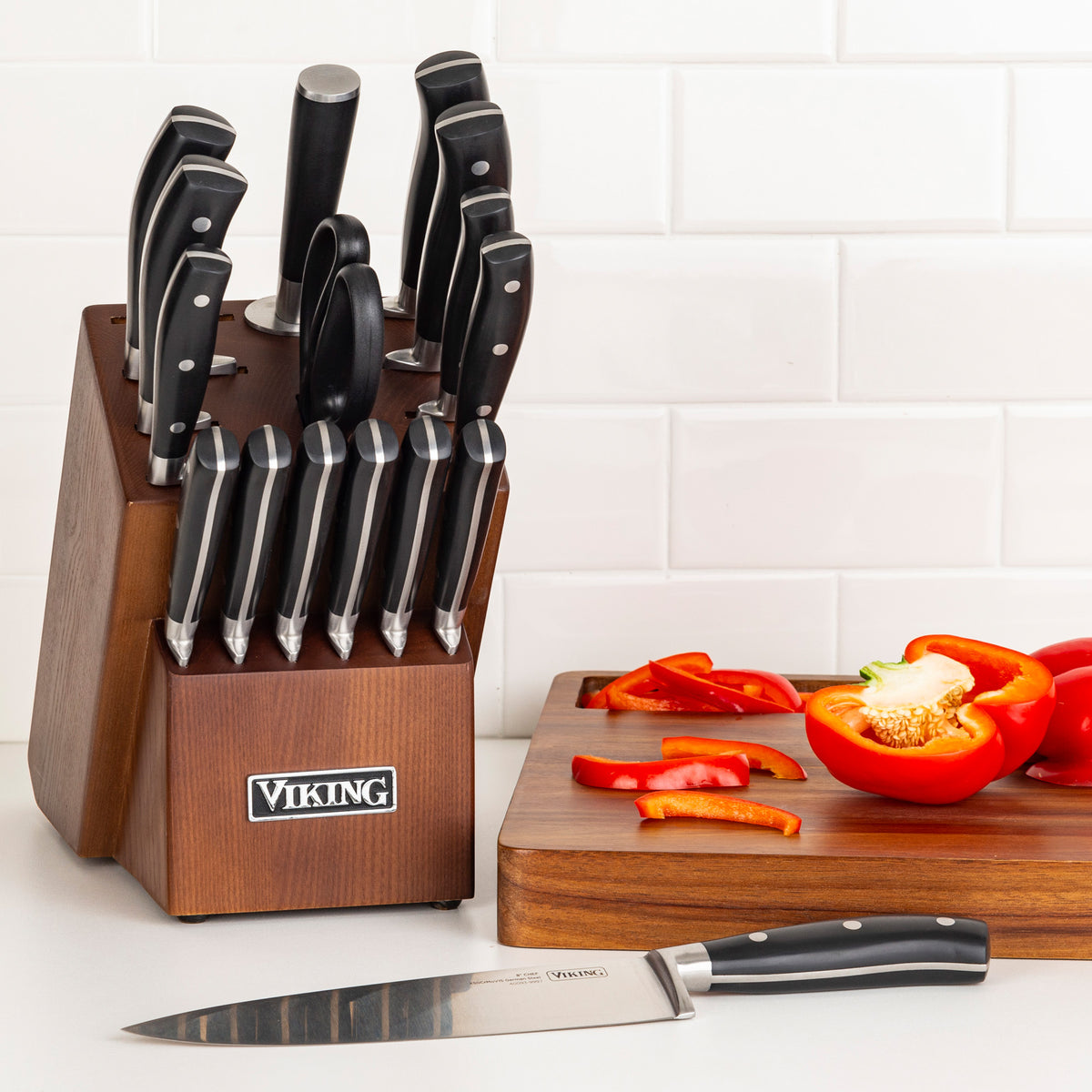 The Viking 17-Piece Cutlery Set with Light Walnut Color Block by Viking Culinary Products, including chef knives of X50CrMoV15 stainless steel and kitchen shears, rests on a counter beside a cutting board with cut and whole red bell peppers against white tiles.