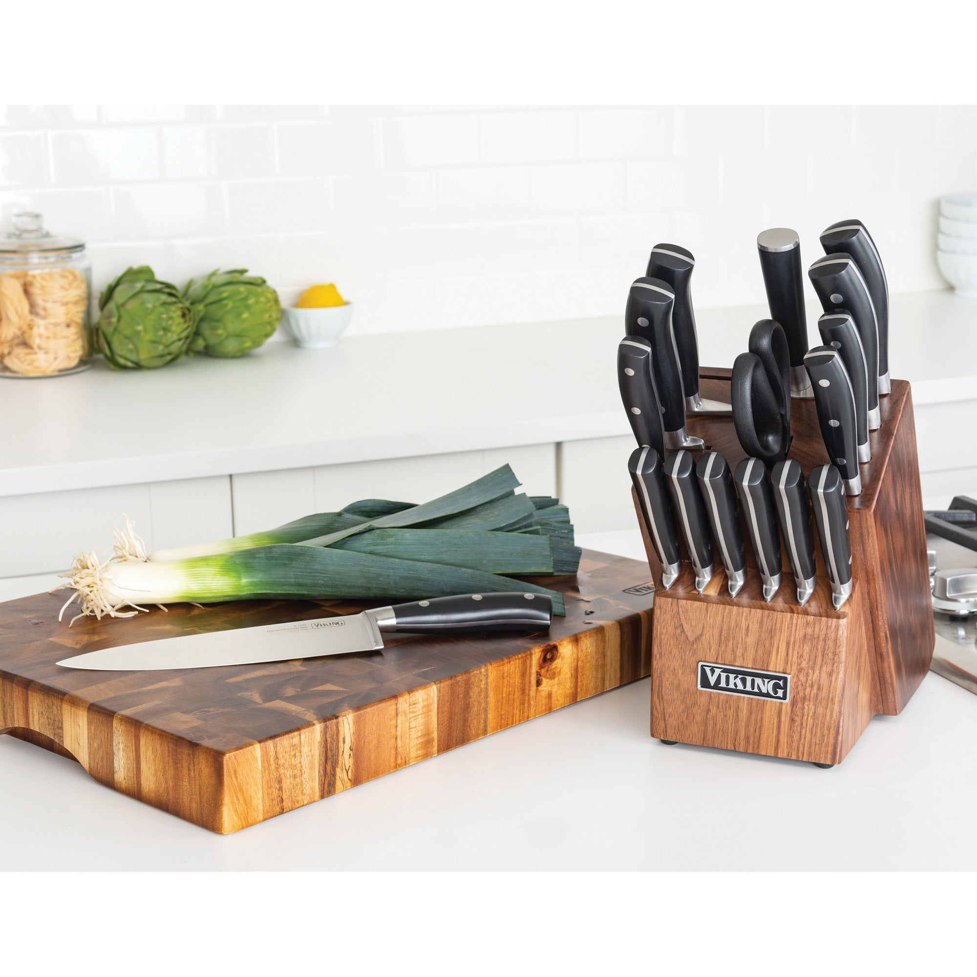 The Viking 17-Piece Cutlery Set with Light Walnut Color Block by Viking Culinary Products rests on the counter near a cutting board topped with a leek, while jars, artichokes, and a lemon add color to the inviting kitchen backdrop.