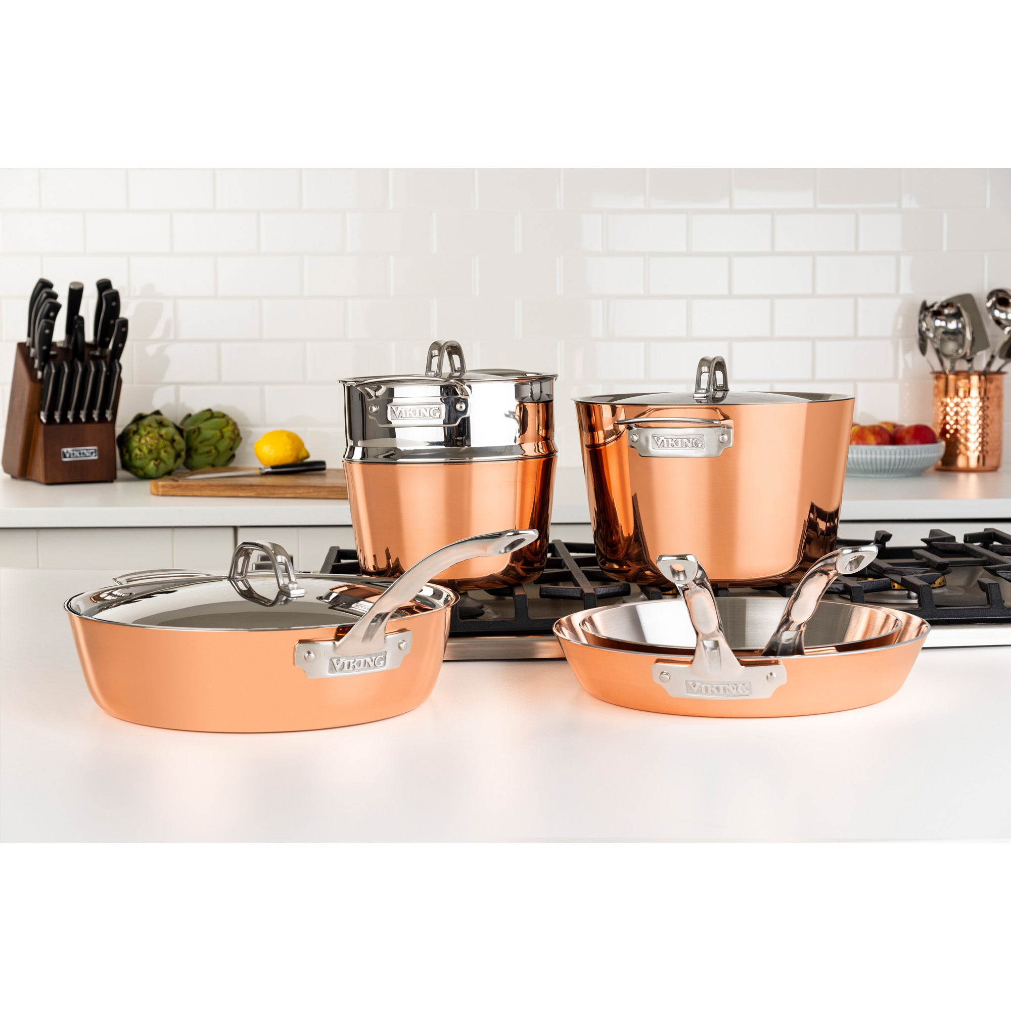 The Viking Contemporary 4-Ply Copper Clad 9-Piece Cookware Set by Viking Culinary Products, featuring induction-capable pots and pans with metal lids, is displayed on a modern kitchen stovetop with a white tile backsplash.