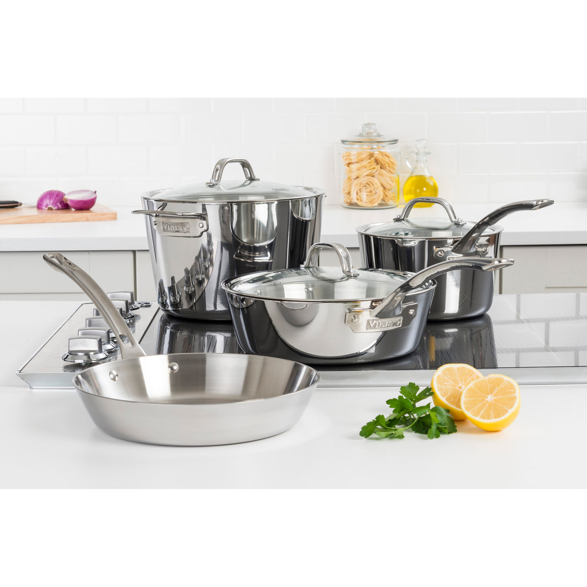 A Viking Culinary Products Contemporary 3-Ply Stainless Steel 7-Piece Cookware Set with glass lids sits on a modern stovetop, surrounded by fresh parsley, a halved lemon, olive oil, and pasta for an inviting kitchen scene.