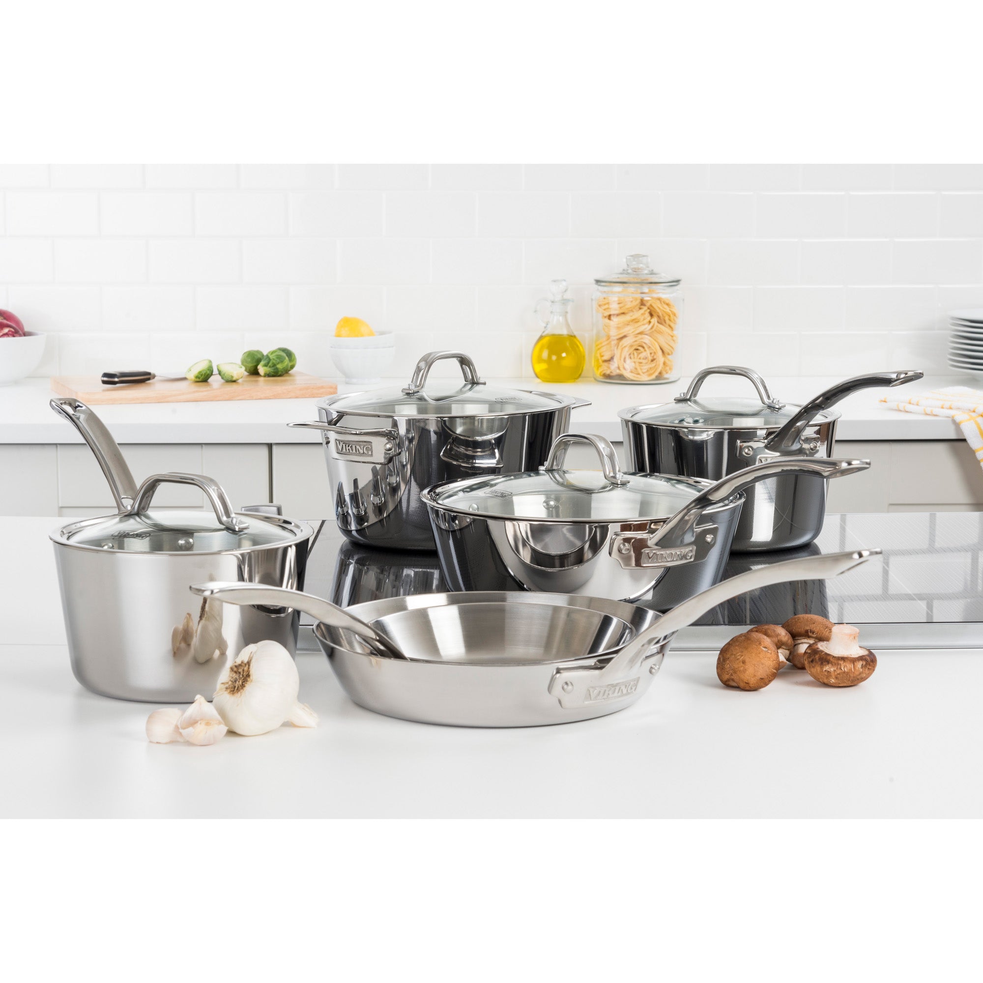 The Viking Contemporary 3-Ply Stainless Steel 10-Piece Cookware Set with Glass Lids by Viking Culinary Products, displayed on a white countertop with garlic, mushrooms, and kitchen items. Includes fry pan, pots, pans with lids. Limited lifetime warranty.