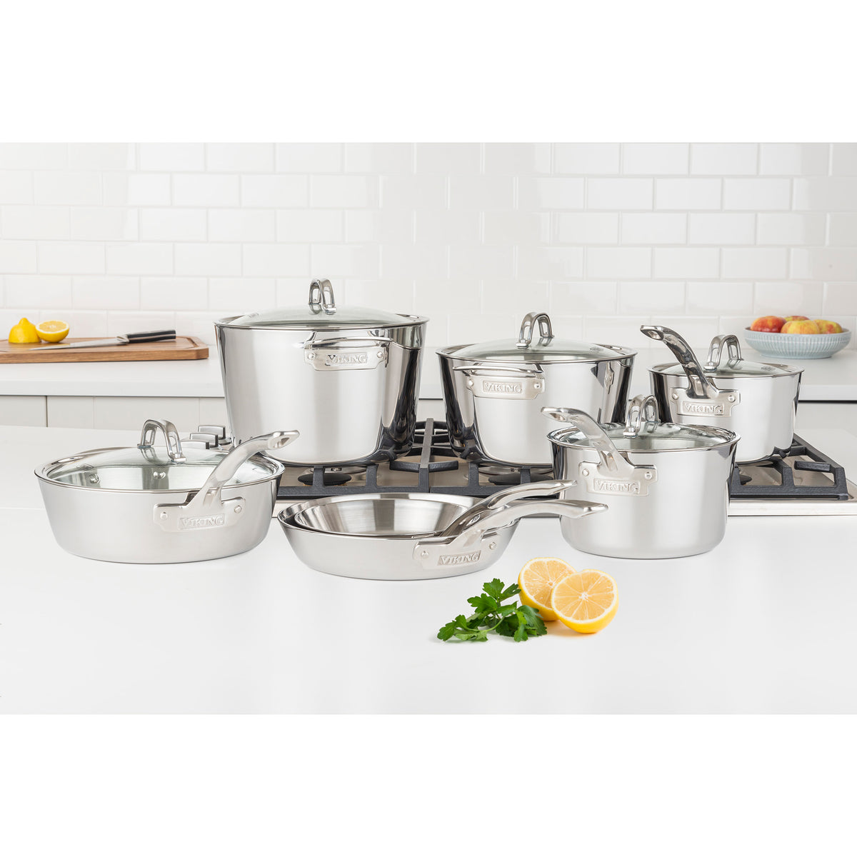 The Viking Culinary Products Contemporary 3-Ply Stainless Steel 12-Piece Cookware Set with Glass Lids is displayed on a stove and countertop in a bright, modern kitchen, surrounded by parsley, a halved lemon, and bowls of fruit.