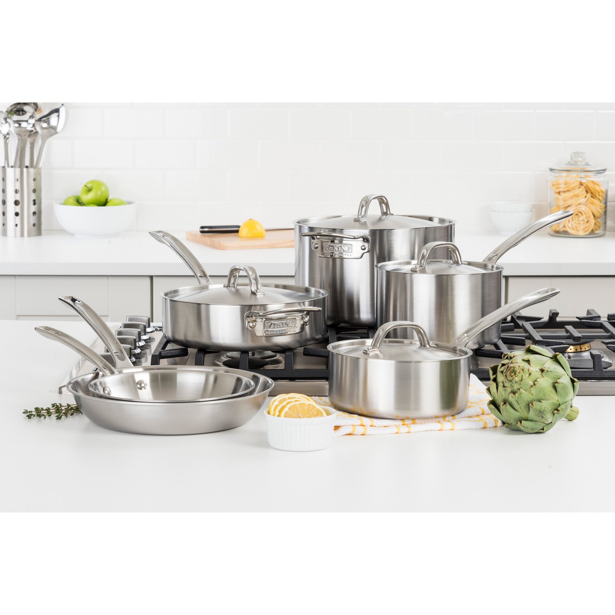 The Viking Professional 5-Ply 10-piece Cookware Set by Viking Culinary Products is displayed on a stove in a modern kitchen, surrounded by a dish towel, artichoke, lemon, and other kitchen items.