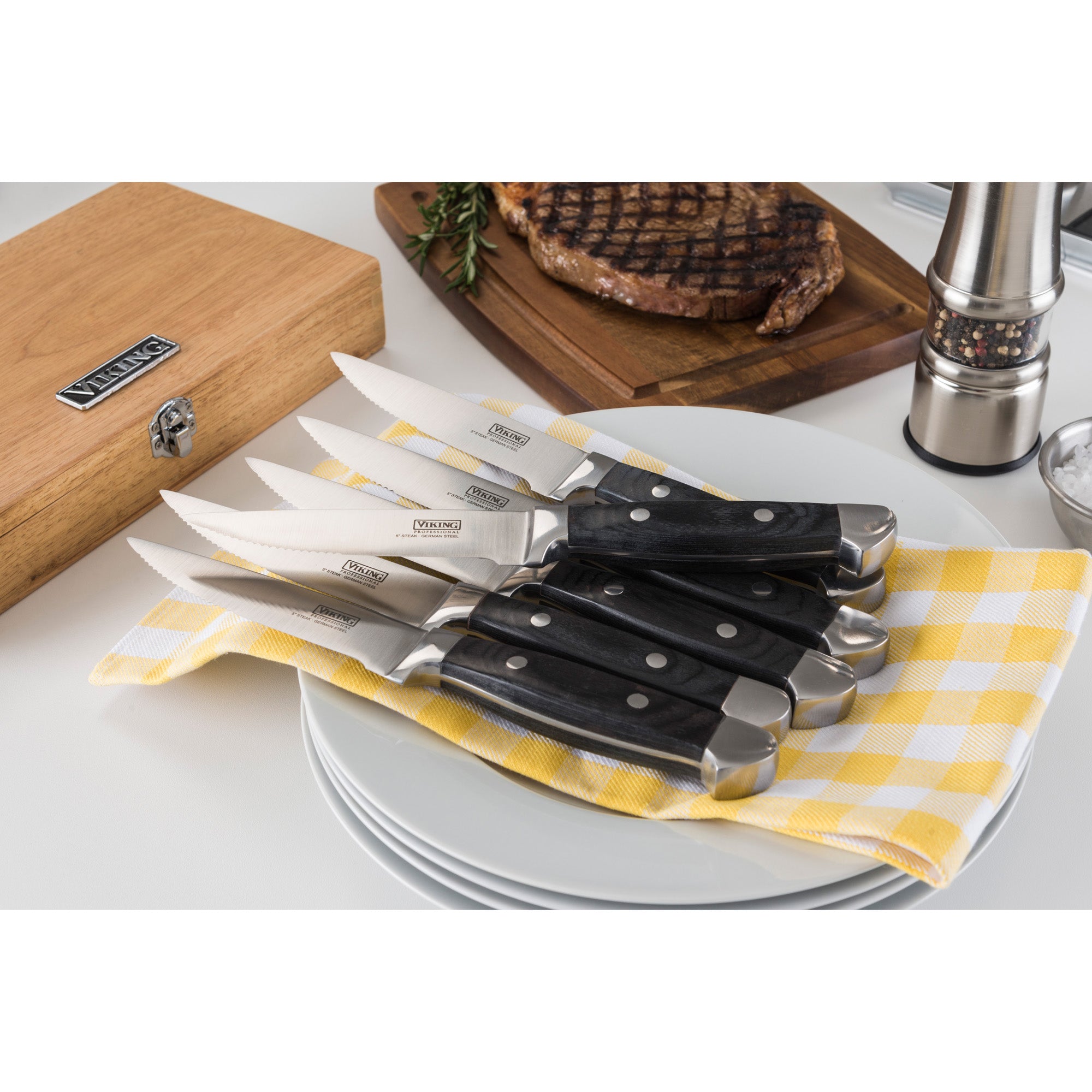 Six Viking Steakhouse Pakka Wood steak knives with black handles from Viking Culinary Products rest on white plates atop a yellow checkered cloth, alongside a wooden gift box, cooked steak, pepper grinder, and rosemary.