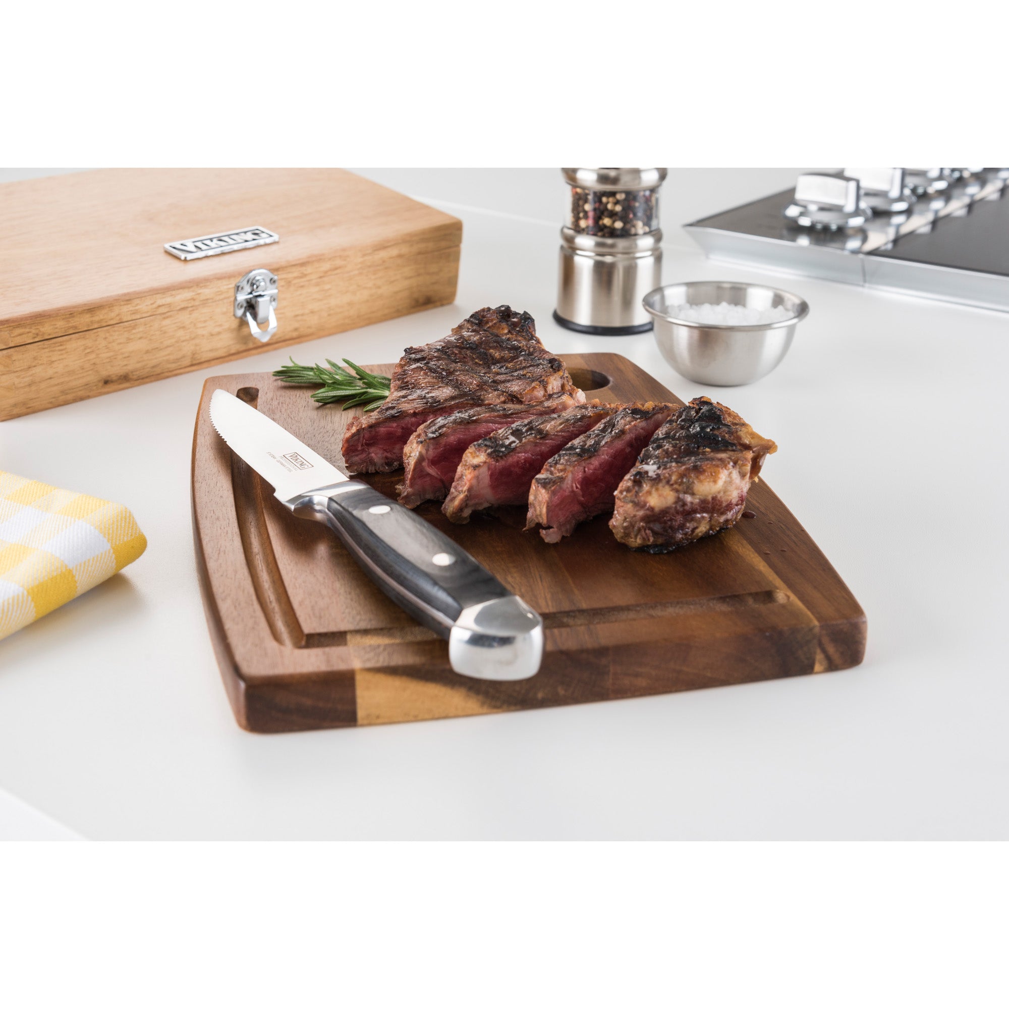 Sliced grilled steak on a wooden cutting board with a Viking Steakhouse Pakka Wood steak knife by Viking Culinary Products. The 6-piece set in black comes with a gift box, shown alongside salt, pepper, and a yellow napkin on a white countertop.