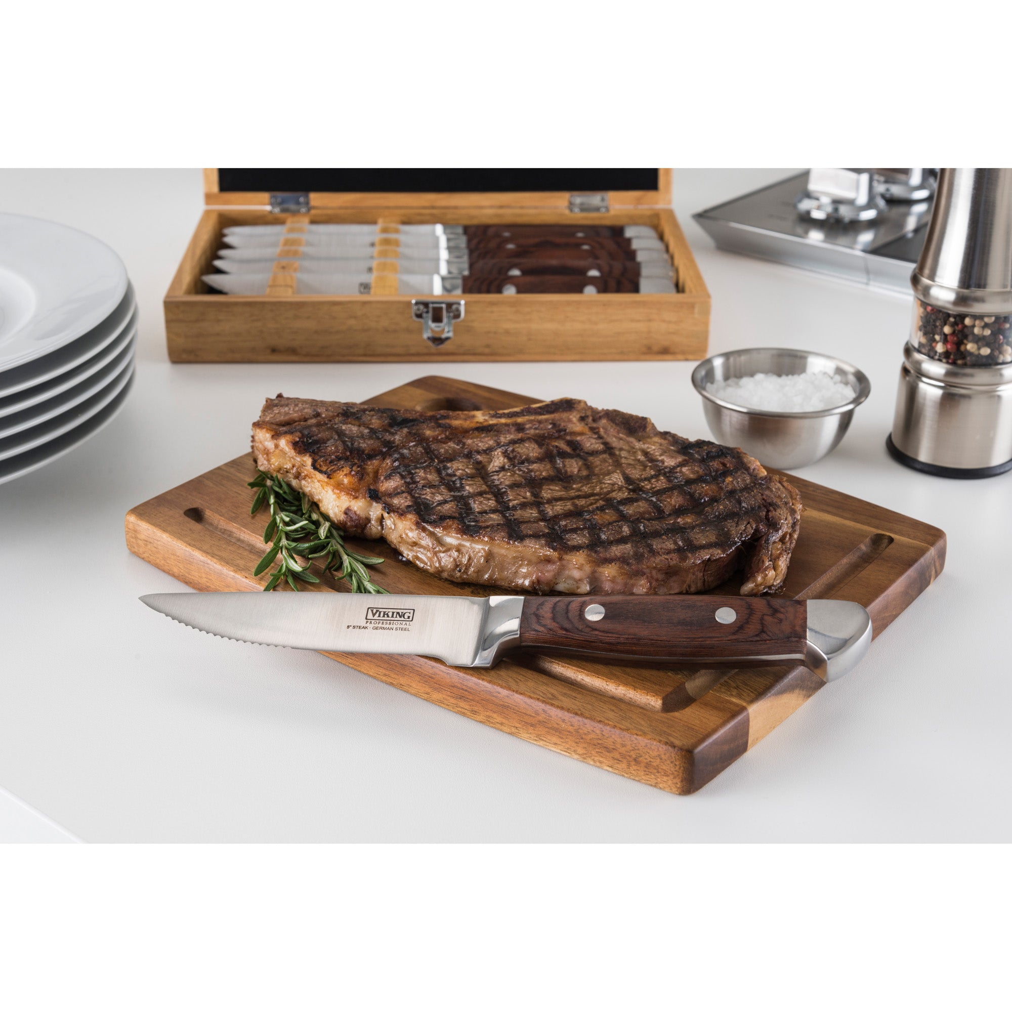 A grilled steak sits on a wooden board with a Viking Steakhouse Pakka Wood Steak Knife by Viking Culinary Products. Behind it are the set’s red gift box, stacked plates, a salt bowl, and a pepper grinder.