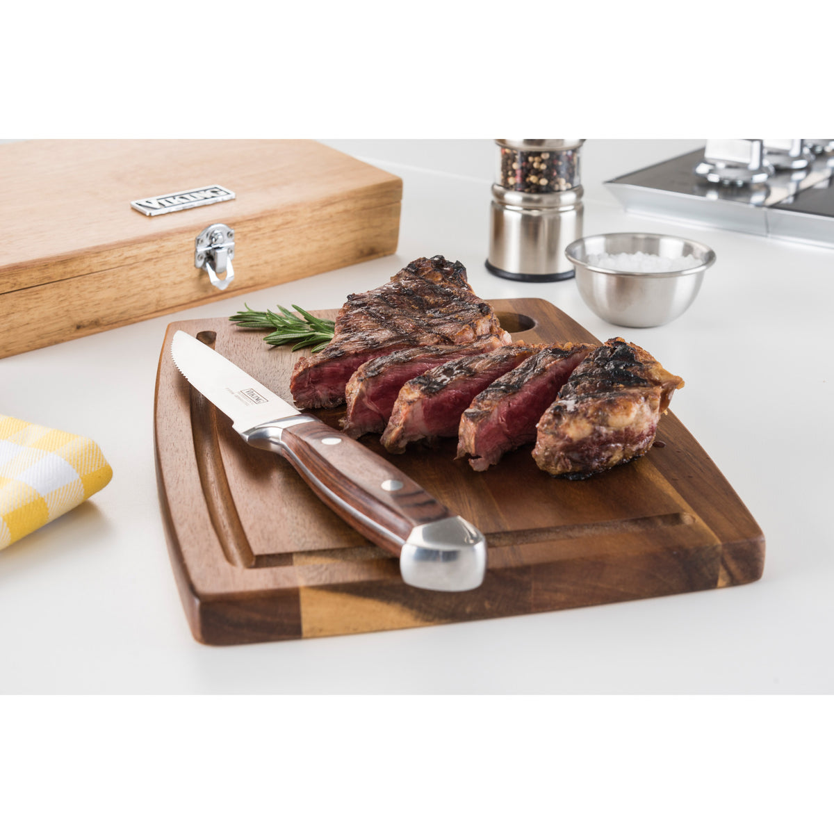 A grilled steak, rosemary sprig, salt bowl, and pepper grinder are arranged with Viking Culinary Products Viking Steakhouse Pakka Wood 6-Piece Steak Knife Set with Gift Box (Red) on a wooden board atop a white kitchen counter.