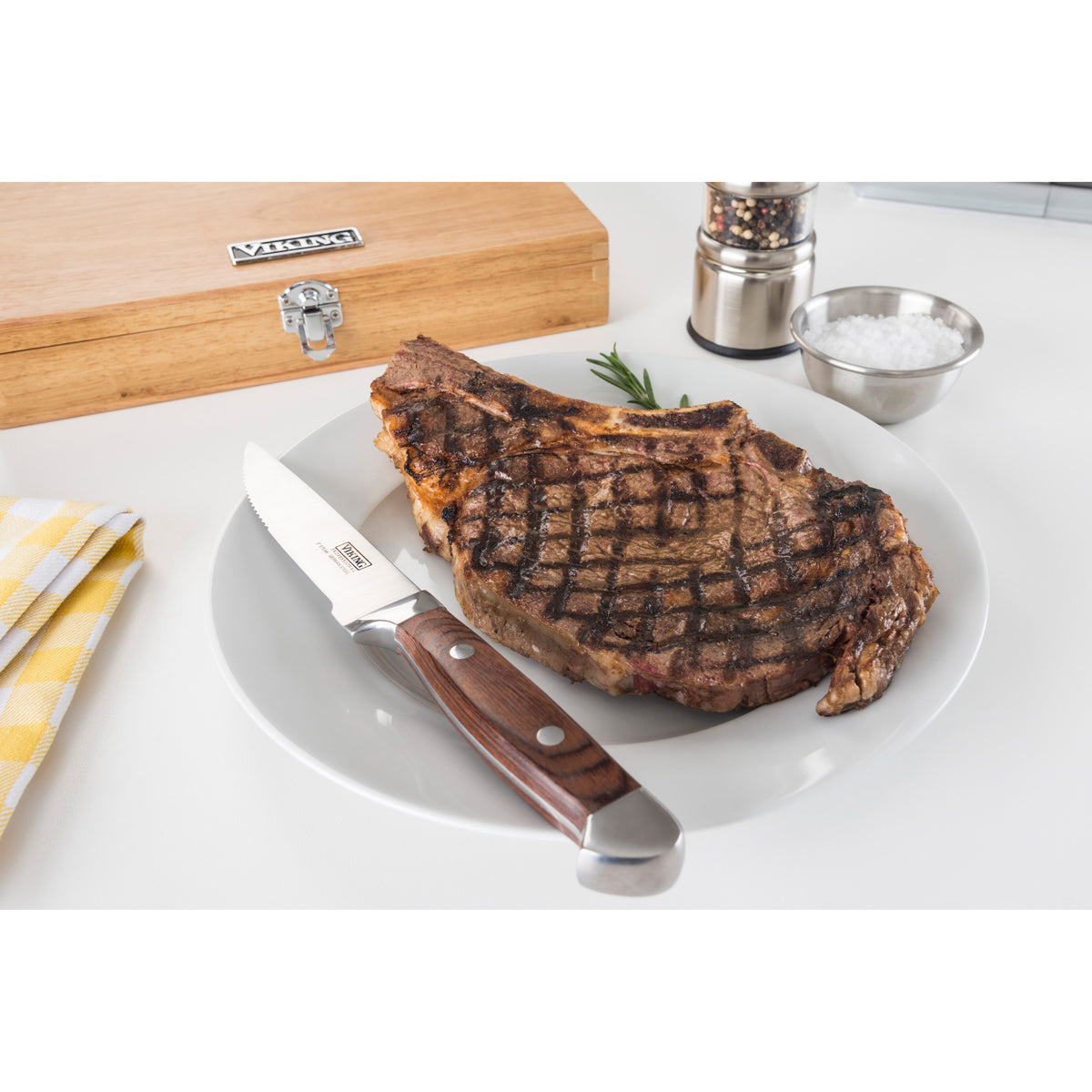 A grilled steak with char marks rests on a white plate, alongside a Viking Steakhouse Pakka Wood Steak Knife by Viking Culinary Products. A red gift box, pepper grinder, bowl of salt, rosemary sprig, and yellow plaid napkin are nearby.