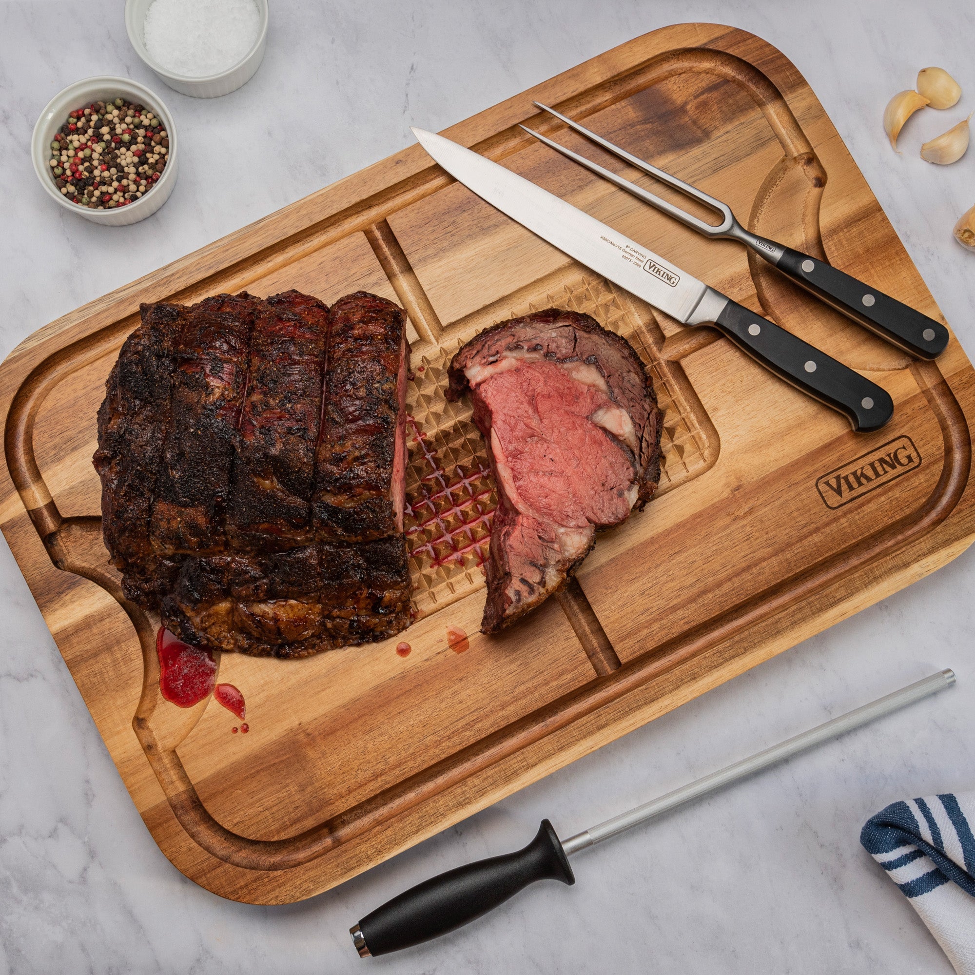 A cooked roast sits on the Viking Acacia Wood Cutting Board, displayed with the 3-Piece Stainless Steel Carving Set by Viking Culinary Products and bowls of salt and peppercorns atop a marble countertop.
