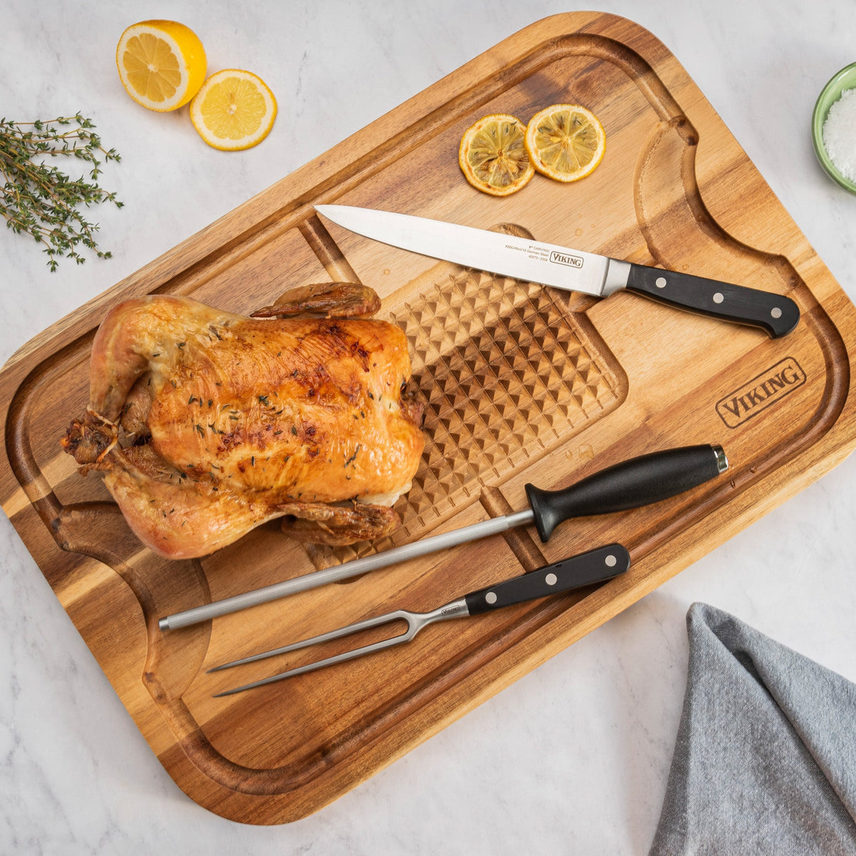 A roasted chicken sits on the Viking Acacia Wood Cutting Board with 3-Piece Stainless Steel Carving Set by Viking Culinary Products. Sliced lemons, fresh herbs, and a folded gray cloth complete the scene.