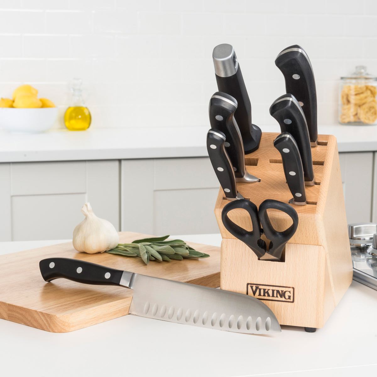 The Viking Professional 9-Slot Beechwood Knife Block by Viking Culinary Products stores knives and scissors neatly on your kitchen counter. Chef’s knife, garlic, and herbs sit on a board nearby, with jars and lemons in the background. Includes lifetime warranty.