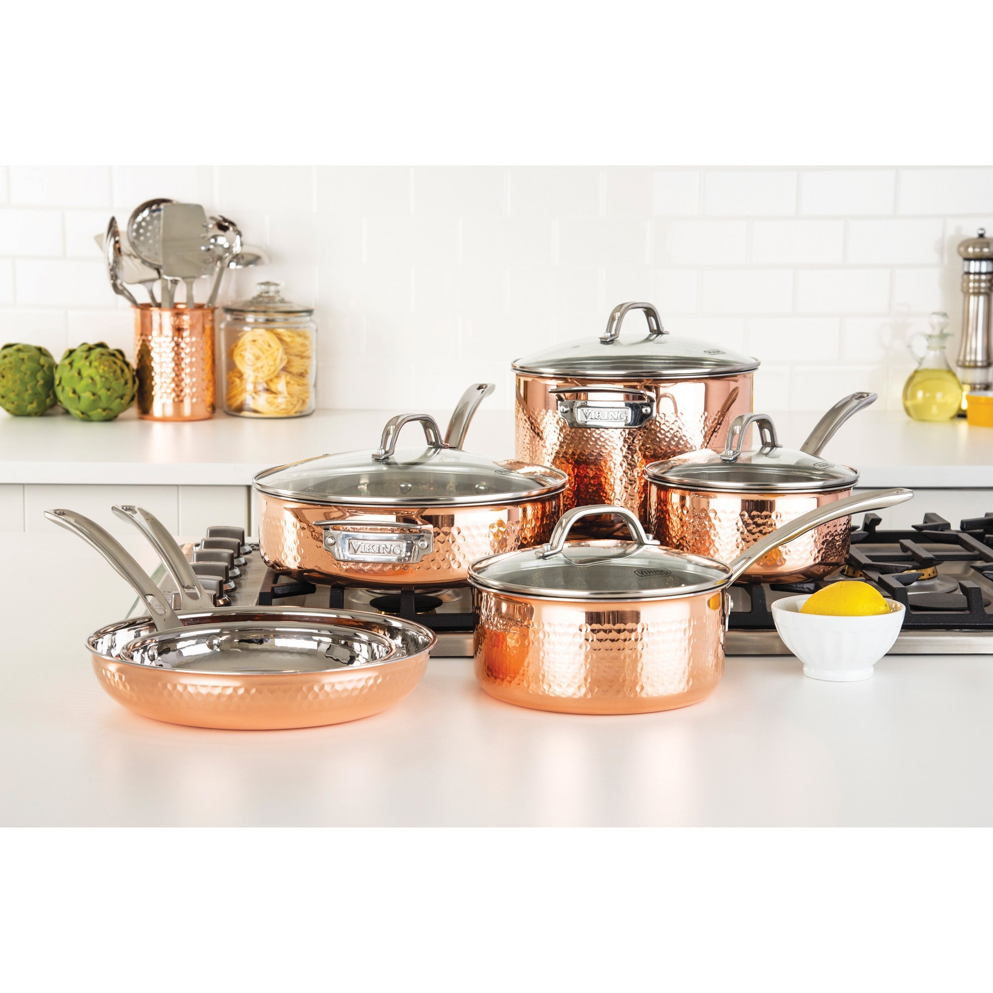 The Viking 3-Ply Hammered Copper Clad 10-Piece Cookware Set with Glass Lids by Viking Culinary Products is displayed on a white kitchen counter and stove, surrounded by utensils, artichokes, pasta jars, and a lemon.